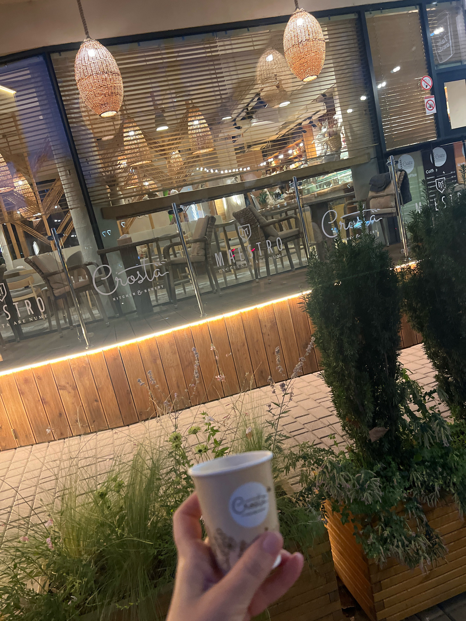 An exterior shot shows a person's hand holding a takeaway coffee cup in front of a restaurant called "Crosta Kitchen & Bakery." The restaurant has a glass facade with wooden blinds and warm interior lighting. In the foreground, there are planters with green foliage and ornamental grasses. The pavement is made of interlocking bricks. The scene is set in an urban environment, likely at dusk or in the evening.