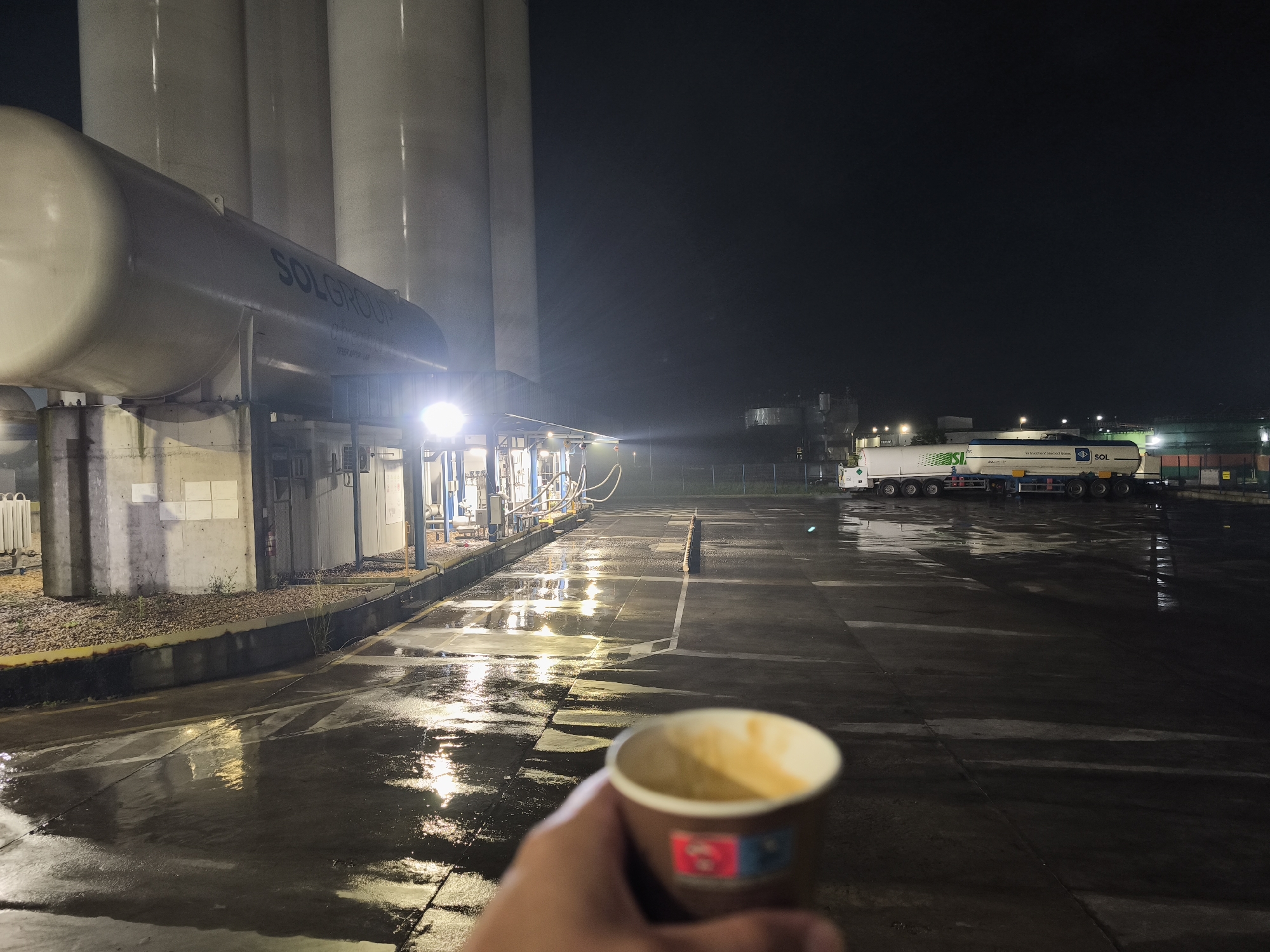 An outdoor, night-time scene at an industrial facility shows a hand holding a disposable cup of beverage in the foreground. The background features large cylindrical storage tanks, one with "SOLGROUP" visible on its side, and industrial piping and equipment illuminated by bright lights. To the right, a tanker truck with "SOL" and other text on its side is parked on a wet, reflective asphalt surface. The ground is wet, suggesting recent rain or a spill, with streaks of light reflecting off it. The sky is dark.