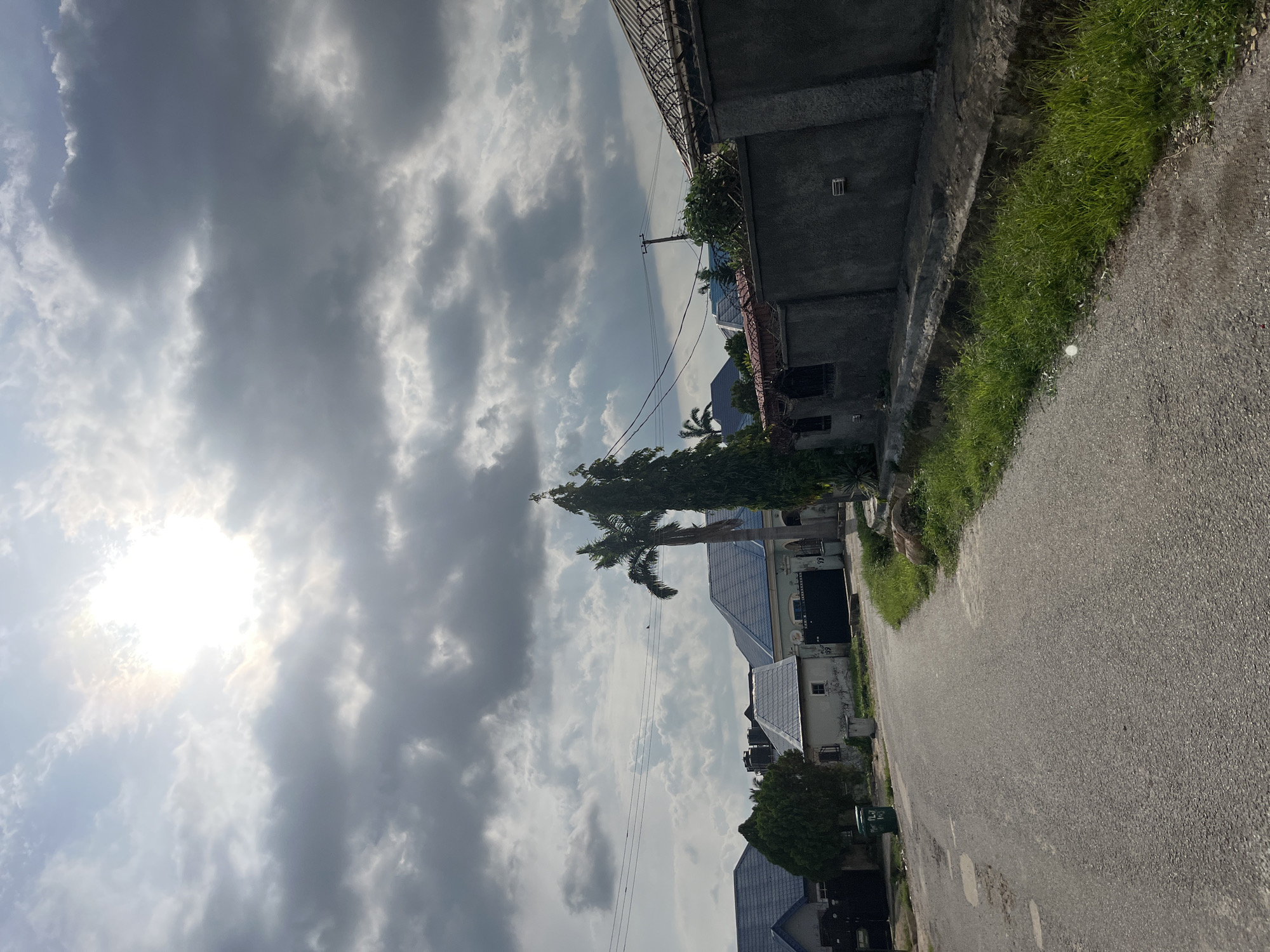 The image depicts a residential street in Abuja, Nigeria, under a partially cloudy sky. The sun is visible through the clouds, casting a bright glow. On the left side of the street, there are houses with blue tiled roofs and some greenery. A tall palm tree stands in the center of the image, alongside other trees. On the right side, a gray concrete wall lines the street, with a drain and grass growing beside it. The asphalt road shows signs of wear and tear with some patching. Overhead power lines are also visible. There are no people or vehicles present in the scene. The overall impression is of a quiet residential area in Abuja.