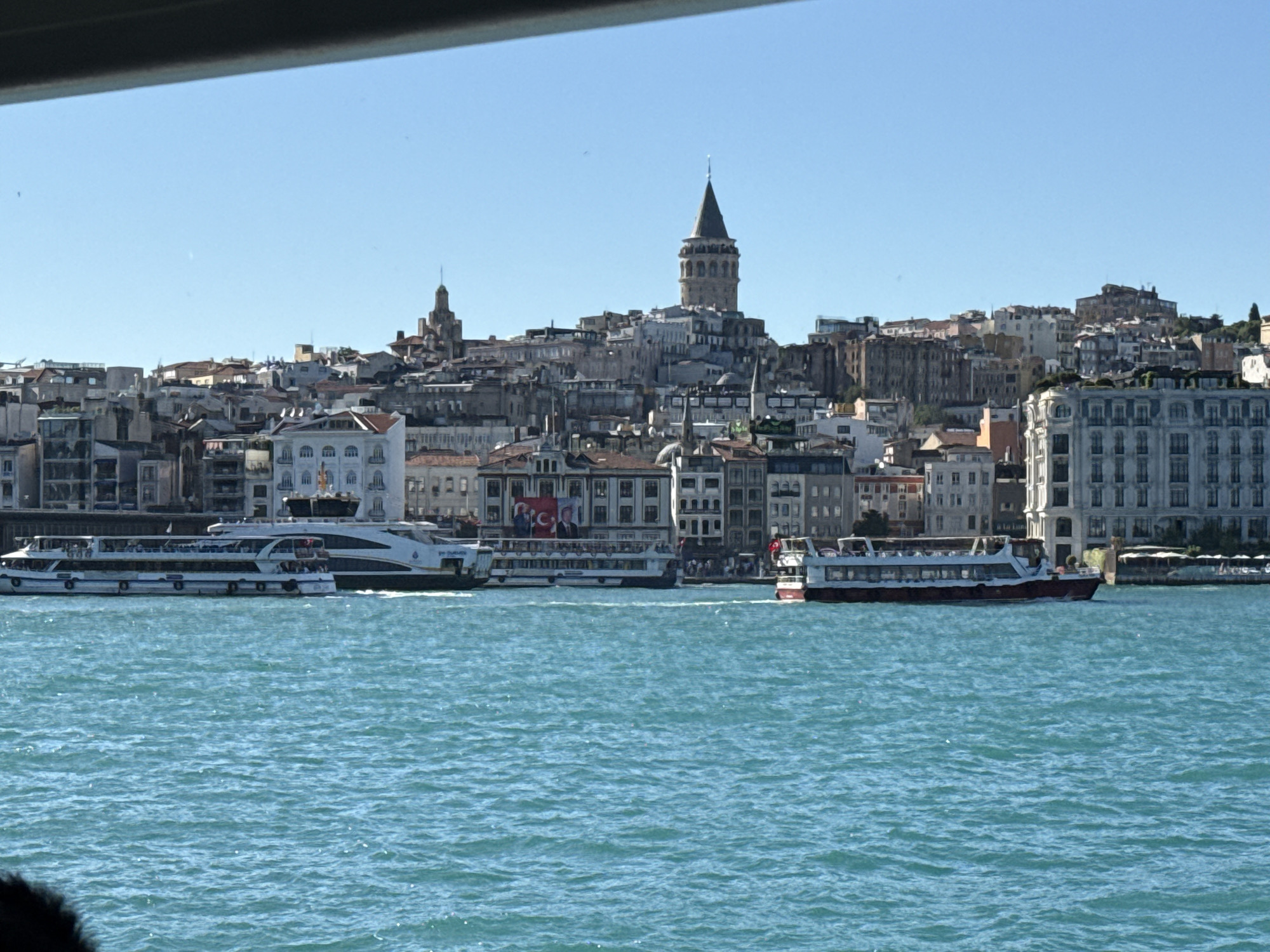 The image depicts a cityscape in Beyoğlu, Turkey, dominated by the iconic Galata Tower, which stands prominently amid a dense collection of buildings. A smaller, steeple-topped structure is situated to the left of the Galata Tower. The architecture is a mix of building styles with visible rooftops and facades.
In the foreground, the turquoise-colored water of the Bosphorus is visible with several passenger ferries navigating the waterway. A series of three ferries appear to be the focus, running parallel to the shore; their color scheme is predominantly white and red. Visible behind and among the ferries are buildings adorned with Turkish flags and portraits, likely of political figures.