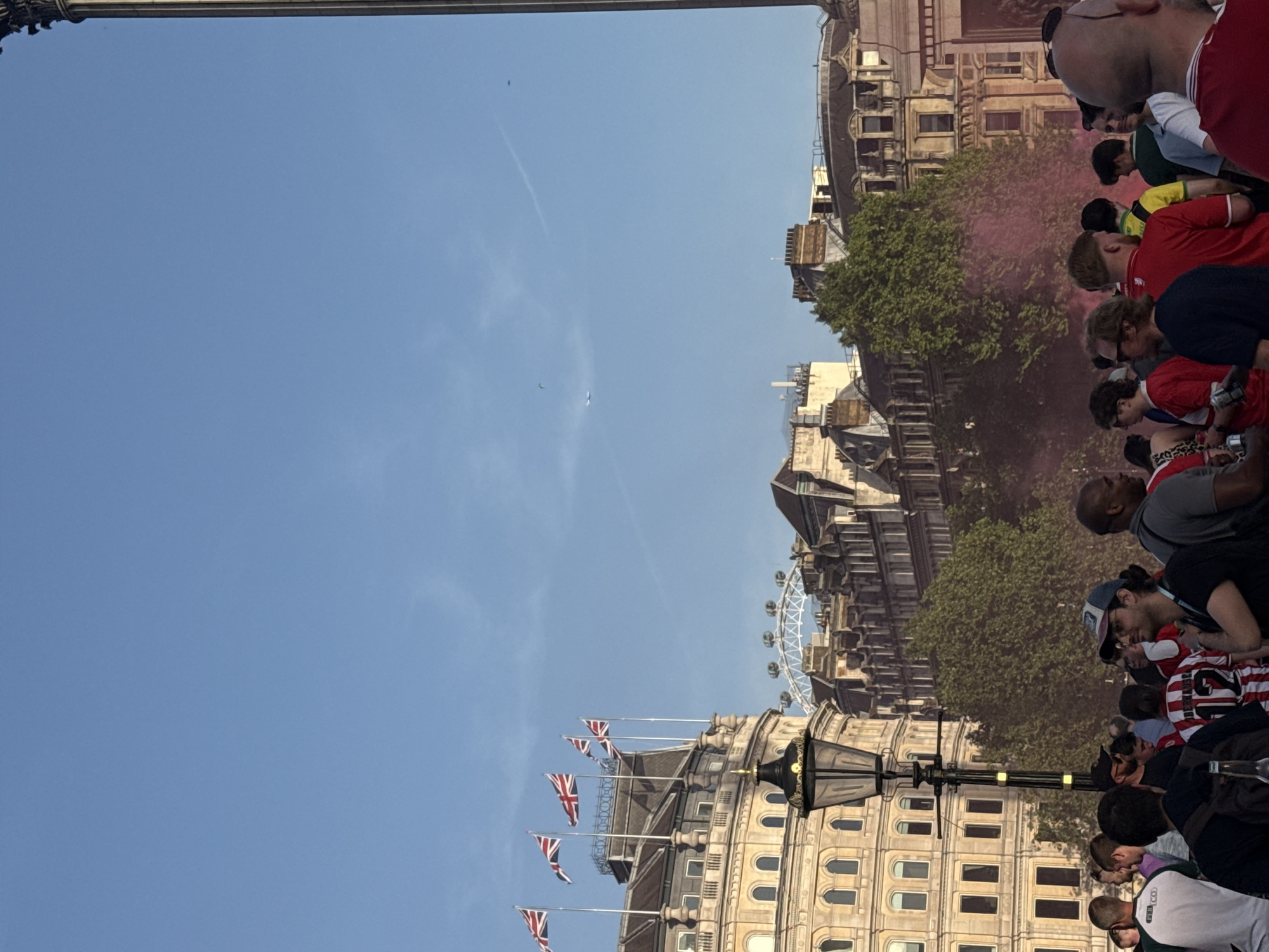 The image depicts a scene in London, United Kingdom. A crowd of people, many wearing red and white sports attire, is gathered along a street lined with buildings. Pink smoke partially obscures the background. Flags are displayed atop one building. The London Eye is visible in the distance. A street lamp is in the foreground. The sky is blue with a few clouds and an airplane.