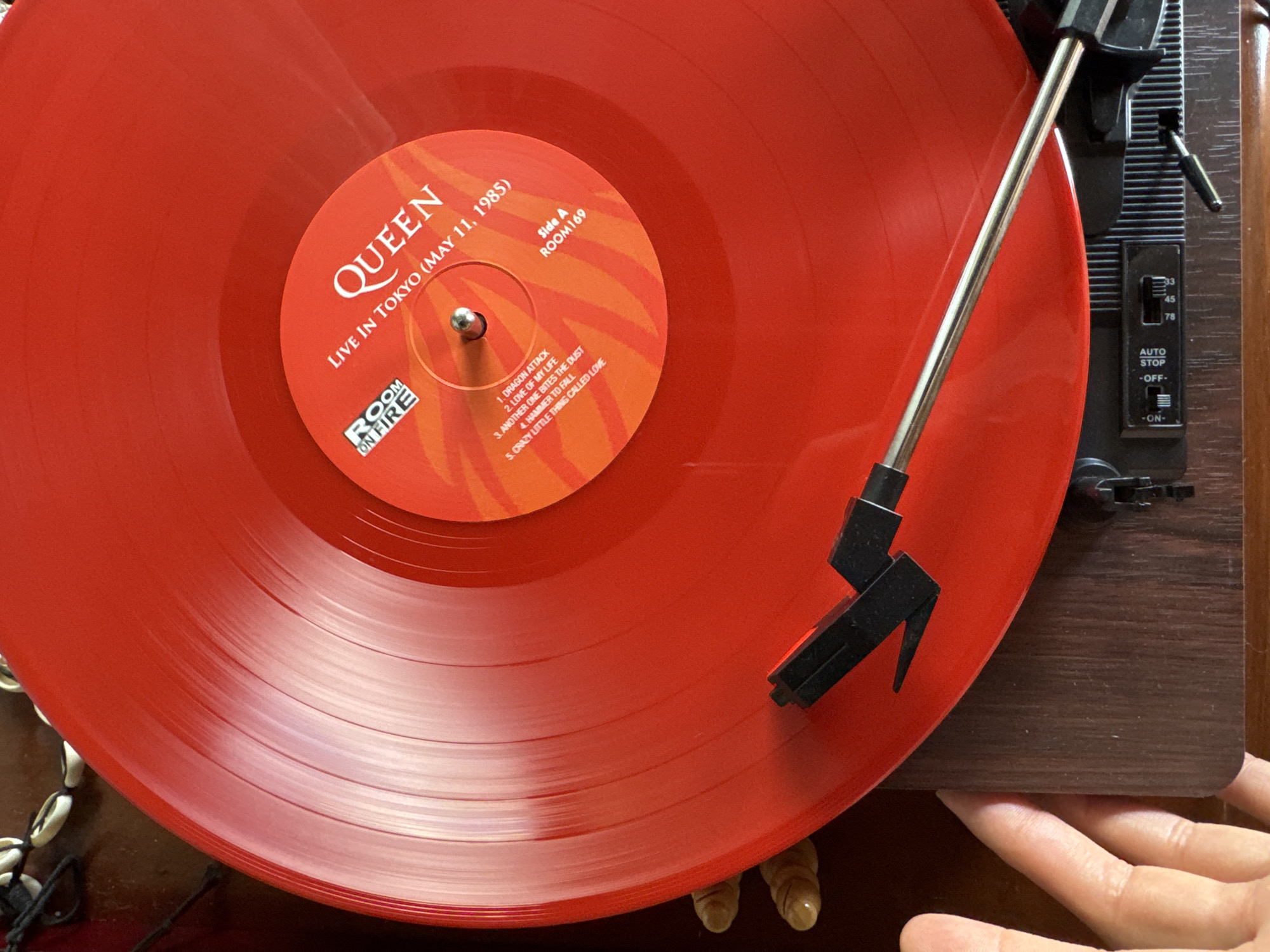 An eye-level, overhead shot shows a red vinyl record of Queen's "Live In Tokyo (May 11, 1985)" on a turntable. The record's label indicates "Side A" and a catalog number "ROOM169," listing the songs "Dragon Attack," "Love Of My Life," "Another One Bites The Dust," "Hammer To Fall," and "Crazy Little Thing Called Love." The turntable's arm and stylus are positioned on the record. The turntable's speed settings (33, 45, 78) and an auto-stop switch are visible. A hand is seen touching the edge of the turntable. The location in Ungasan, Indonesia is not discernible from the image.