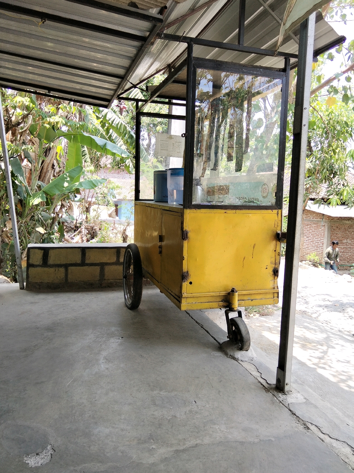 This is a full outdoor shot of a yellow food cart parked under a metal roofed shelter. The cart is made of metal with glass panels on the front and top, displaying a menu. It has two wheels, one visible on the right side, and is positioned on a concrete ground. Behind the cart, there are banana trees and a brick building. In the background, a person is seen bending down near the building. The scene appears to be in a rural or suburban area of Jenangan, Indonesia, possibly a street-side vendor location.