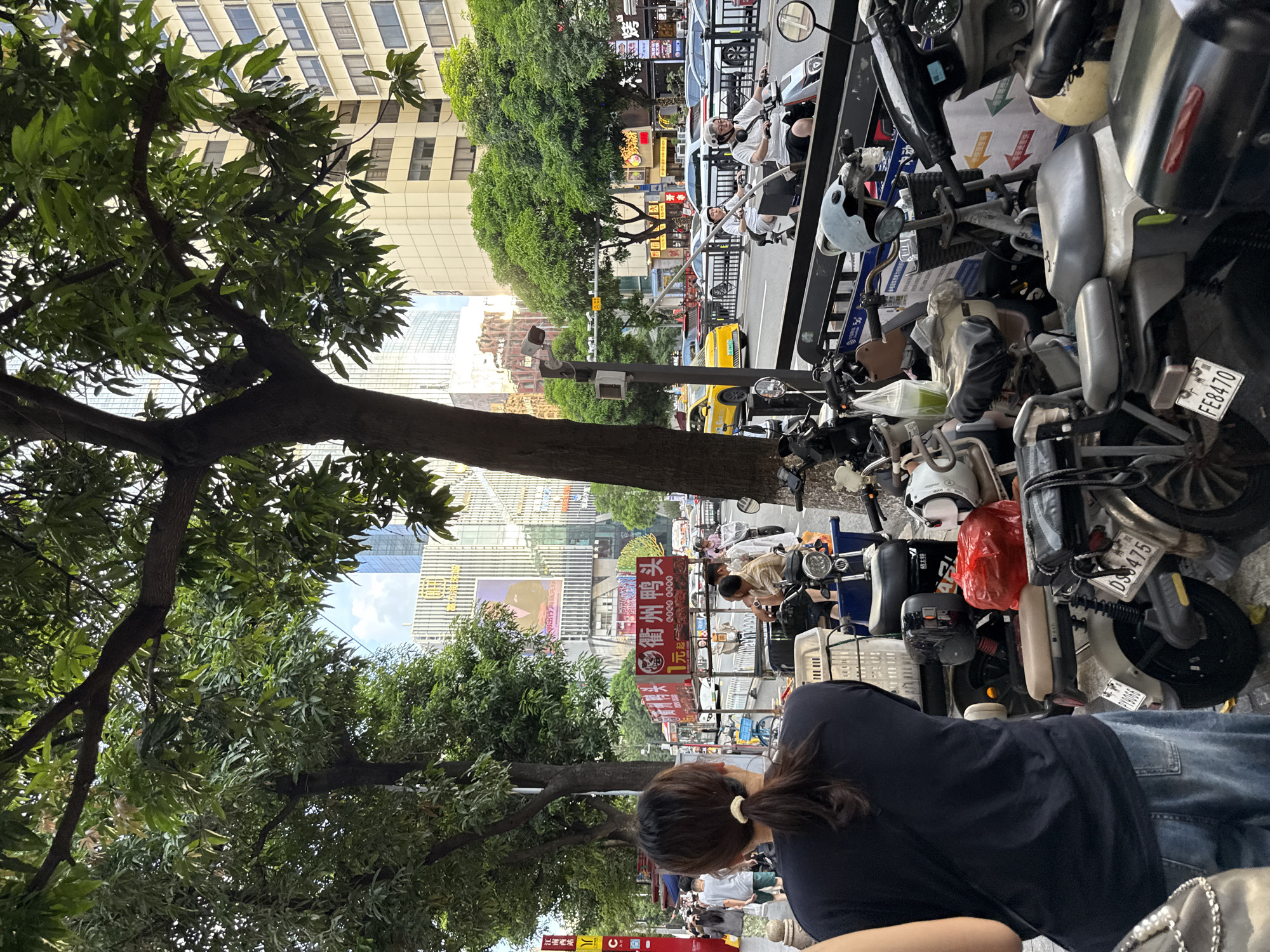 This image captures a street scene in Haizhu District, China, likely during the daytime given the illumination. The foreground is dominated by a collection of parked electric scooters and motorcycles, with some displaying visible license plates such as "DS475" and "FE8470". A woman with her back to the camera, wearing a dark t-shirt and blue jeans, stands amongst these vehicles. Behind her, several other individuals are visible, some walking on the sidewalk and others seated on scooters. In the mid-ground, a street-side food stall with red signage is present, advertising what appears to be duck. Beyond the stalls, traffic moves along a busy road, with cars and a yellow taxi visible. The background features tall, modern buildings, characteristic of an urban cityscape. Lush green trees line the sidewalk, providing shade. Overhead, a clear sky with some clouds is visible. The overall impression is of a bustling urban environment with a focus on pedestrian and small vehicle traffic, typical of a commercial area in a Chinese city.