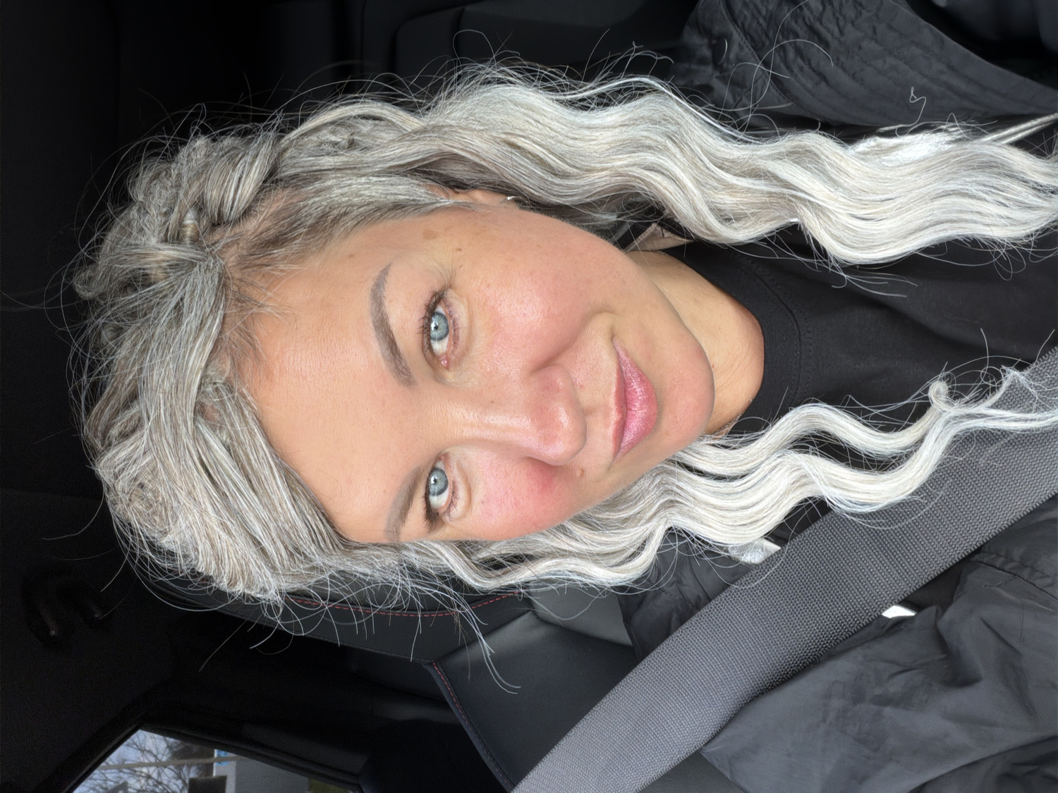 The image displays a close-up portrait of a woman, rotated clockwise by 90 degrees, so she appears to be lying down. She has long, voluminous, wavy silver-gray hair, with subtle darker roots visible near her scalp. A small, delicate braid is present on the left side of her head, near her temple. Her piercing blue eyes look directly at the camera, and she has a soft, closed-mouth smile, with her lips tinted a light pink. Her complexion is fair.
She is wearing a black top or jacket. A gray seatbelt is visible diagonally across her chest, suggesting she is inside a vehicle. The background reveals the dark interior of a car, with black upholstery and subtle red stitching visible on what appears to be a seat. Through a window in the bottom left corner, tree branches and what might be a distant building under an overcast sky can be seen, indicating an outdoor, daytime setting. The woman's expression conveys a pleasant and relaxed demeanor. No visible text is present in the image.