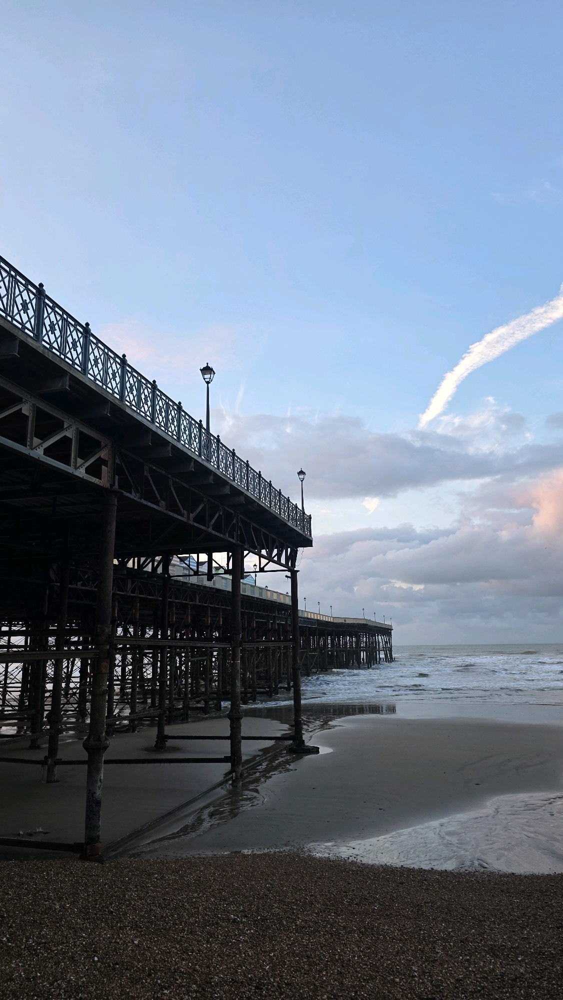 The image shows a view of the pier at St. Leonards-on-Sea, United Kingdom. The pier, constructed of dark metal, extends out over the water. The pilings supporting the pier are visible on the wet sand below. The beach is composed of pebbles in the foreground and transitions to sand closer to the water. Gentle waves lap against the shore. The sky is a mix of blue and grey clouds, with a distinct contrail visible in the upper right portion of the frame. Two lampposts with traditional lantern-style fixtures are positioned along the pier. The scene appears to be captured during either sunrise or sunset, given the soft lighting and muted colors. There are no people or animals visible in the image.