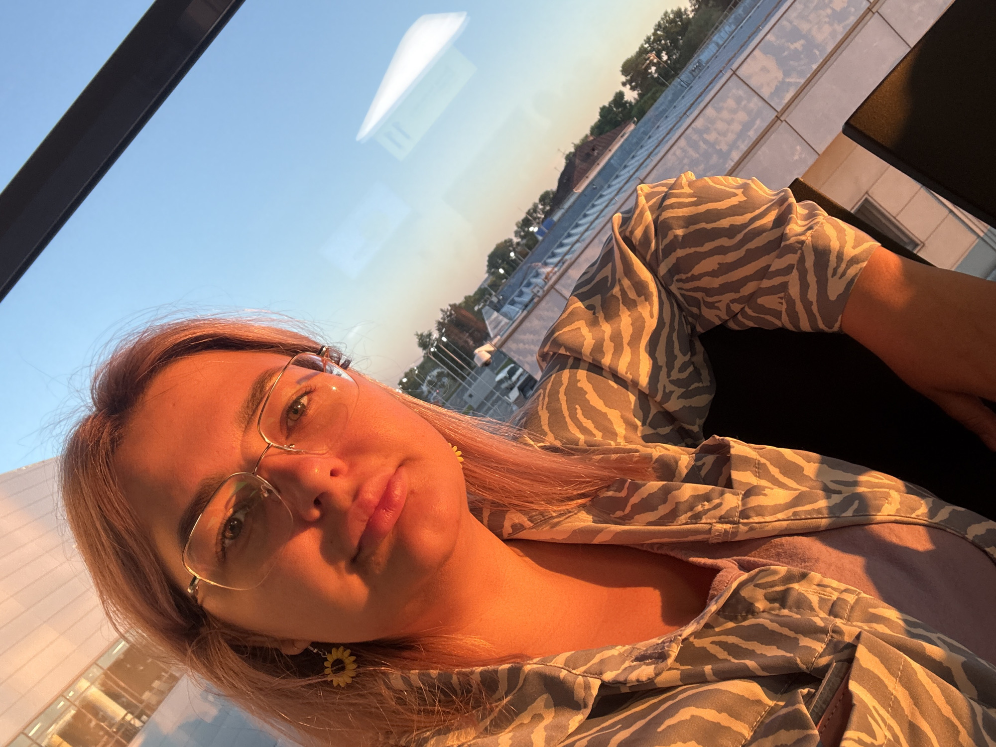 The image shows a close-up, selfie-style shot of a woman wearing glasses and a patterned shirt. The setting appears to be indoors, near a window, with the warm golden light of either sunrise or sunset casting a strong glow on the woman's face and upper body. Outside the window, a clear blue sky is visible, along with reflections of buildings and trees. The woman has blonde hair and is wearing sunflower-shaped earrings. Her clothing has a zebra-like pattern. The overall impression is that of a personal moment captured during golden hour, likely in an urban environment. The image does not contain enough information to confirm or deny its location in Khabarovsk, Russia.