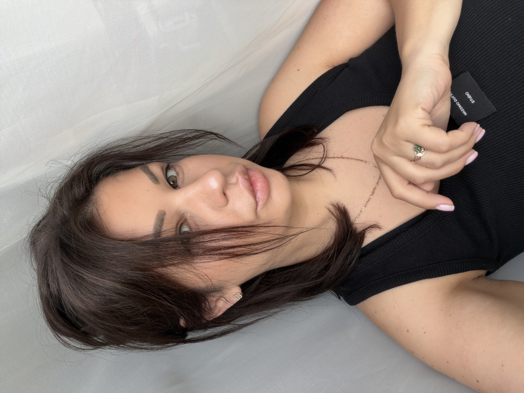 In a medium shot, a young woman with dark brown hair, styled in a messy updo with some strands falling across her face, poses for a selfie against a white, textured curtain backdrop. She is wearing a black, ribbed tank top and a delicate rose gold necklace. Her left hand rests on her chest, drawing attention to a rose gold ring with green stones. A small black tag with white text, possibly a brand label, is visible tucked into the neckline of her top. The lighting is soft and diffused, highlighting her features and the textures of the fabric. There is no discernible indication of the city of Sochi, Russia, or any specific urban environment.
