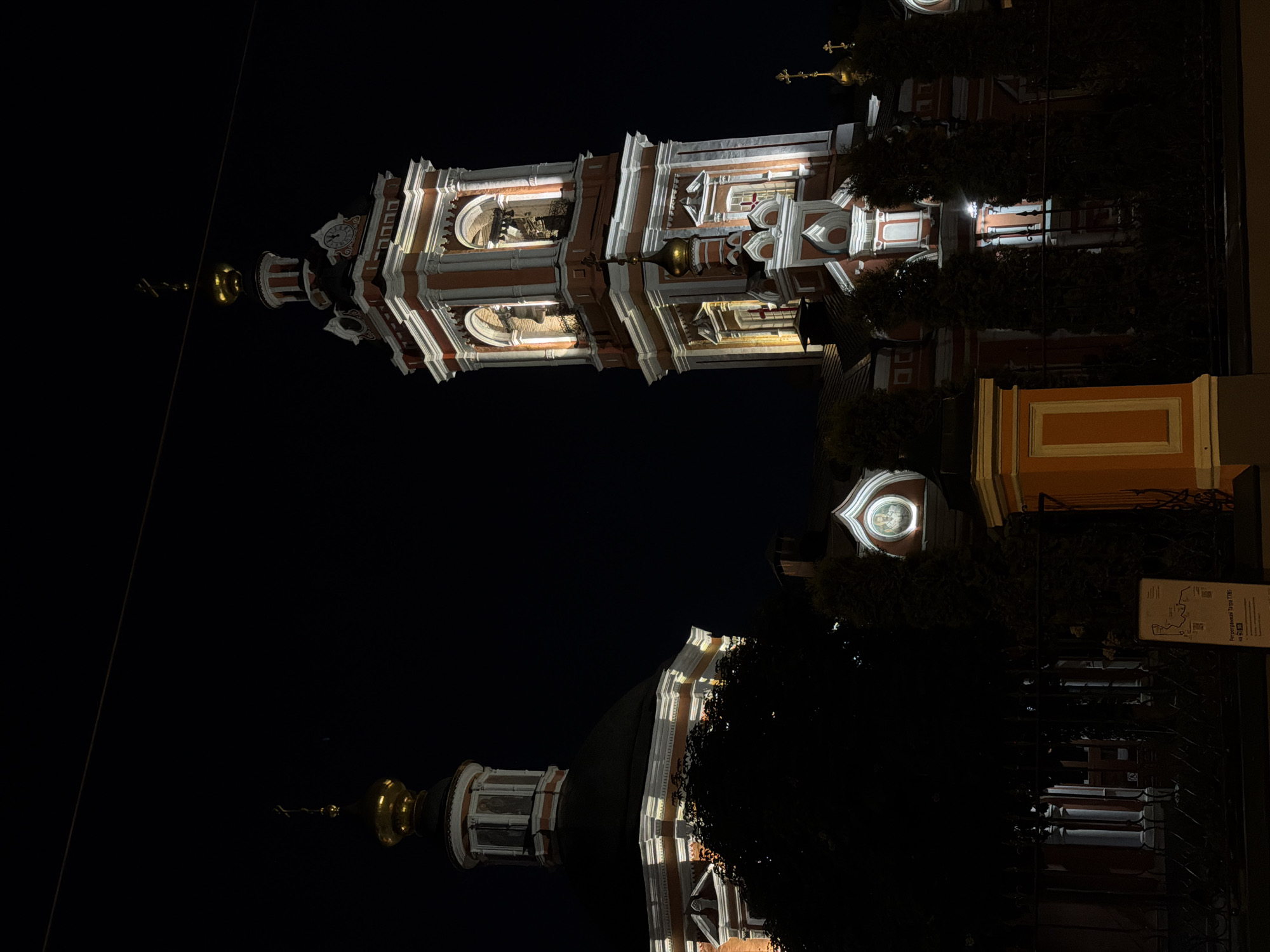 The image shows a nighttime view of a church in Moscow, Russia, specifically depicting the bell tower and adjacent structures illuminated against a dark sky. The architectural style is characterized by red and white detailing, with gilded domes visible on the top left and bottom center of the frame. Parts of the buildings are covered in dark green foliage, obscuring some features. A sign with text and a map is partially visible in the lower right corner.