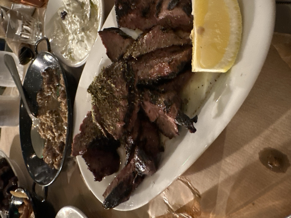 This image captures a close-up view of a meal served indoors, likely at a restaurant in Nea Erithrea, Greece. The primary subject is a white oval plate laden with slices of dark, grilled meat, possibly lamb or beef, generously seasoned with green herbs like oregano or thyme. A bright yellow lemon wedge rests on the side of the plate, indicating it's meant to be squeezed over the meat.
Adjacent to the main plate, there's a small white bowl filled with a creamy, white dip, characteristic of tzatziki, garnished with a few dark specks, possibly olives or spices. To its left, a small black pan or bowl contains a textured, brownish dish, potentially a stew or a mashed legume, with a spoon resting in it. Other blurry food items are visible in the background, suggesting a spread of dishes.
The setting appears to be a dimly lit dining area, with warm artificial light casting shadows. The table is covered with a plain brown paper or tablecloth. There are no people visible, focusing the attention entirely on the food. The overall impression is that of a traditional Greek meal, ready to be enjoyed. No text is visible in the image.