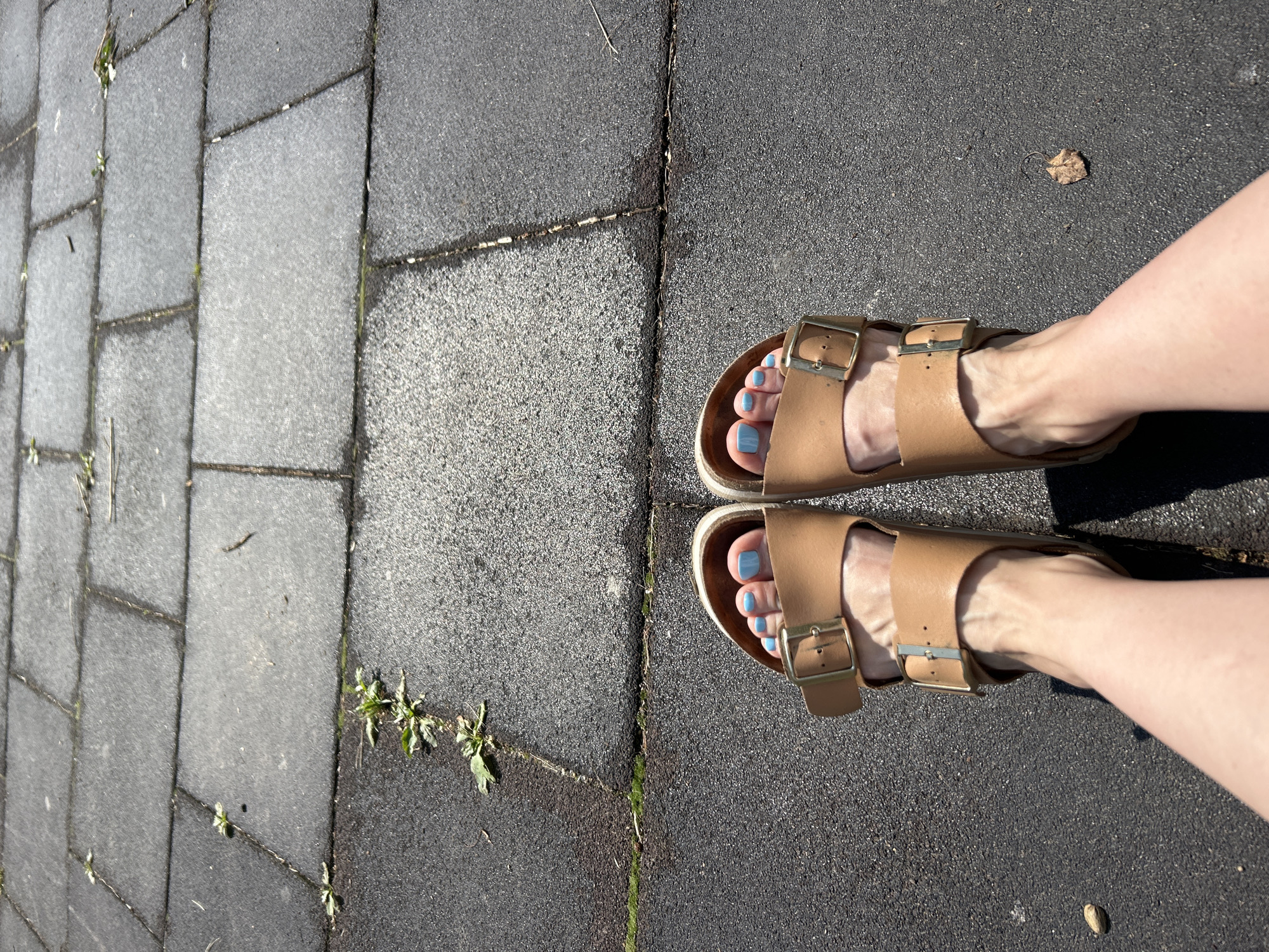 The image shows a pair of light brown Birkenstock sandals with gold buckles worn by a person standing on a paved surface. The pavement is composed of large, rectangular dark gray tiles with visible seams and some small green weeds growing in the cracks. The feet are adorned with light blue nail polish. The lighting suggests an outdoor environment under direct sunlight, casting shadows on the ground. The overall scene appears to be a casual outdoor setting. The location is Chaikovsky, Russia.