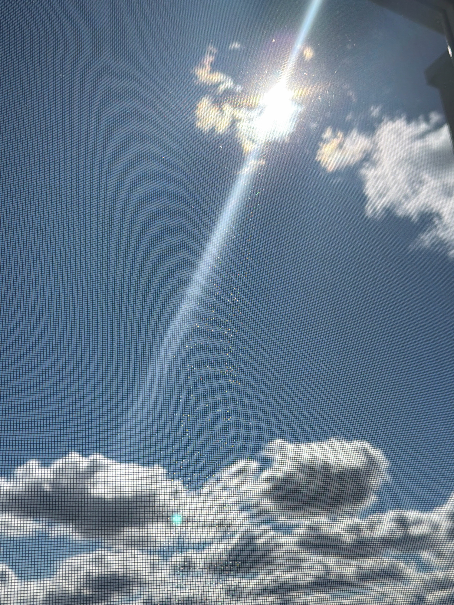 The image depicts a bright, sunny sky filled with white, fluffy clouds, viewed through a window screen. The sun is visible in the upper right quadrant, casting a strong glare and a ray of light that diagonally crosses the frame. Dust particles are visible along the sun's ray, creating a shimmering effect. The bottom portion of the image is dominated by a layer of clouds, with more sparse clouds scattered across the blue sky. The view appears to be from an elevated position, looking upwards. The location is identified as Pavlodar, Kazakhstan.