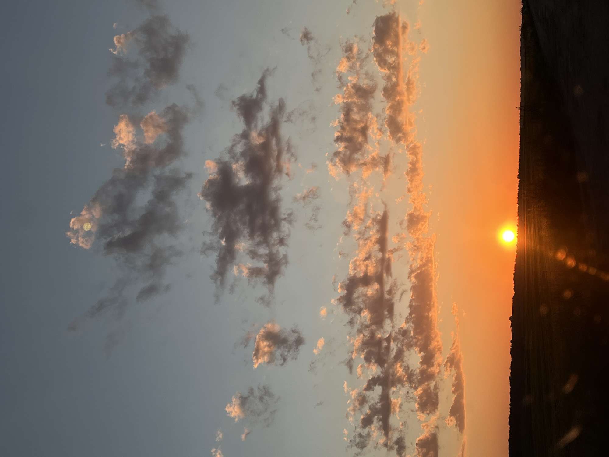 This is a vertical, eye-level, medium shot photograph of a sunset over a rural landscape, presumably near Skomorokhy, Ukraine. The sky dominates the upper two-thirds of the frame, transitioning from a pale blue at the top to a vibrant orange near the horizon. Scattered clouds are present throughout the sky. Some clouds are dark gray and appear heavy, while others are illuminated by the setting sun, glowing with shades of pink and orange. The sun itself is a bright, orange orb low on the horizon, casting a golden light across the scene and creating a lens flare effect in the lower portion of the image. A dark, silhouetted treeline forms the horizon line, separating the sky from the land. The foreground consists of dark, out-of-focus land, likely fields, and a dirt path on the right side of the frame. No people or animals are visible in the image. The overall mood is serene and peaceful, characteristic of a sunset.