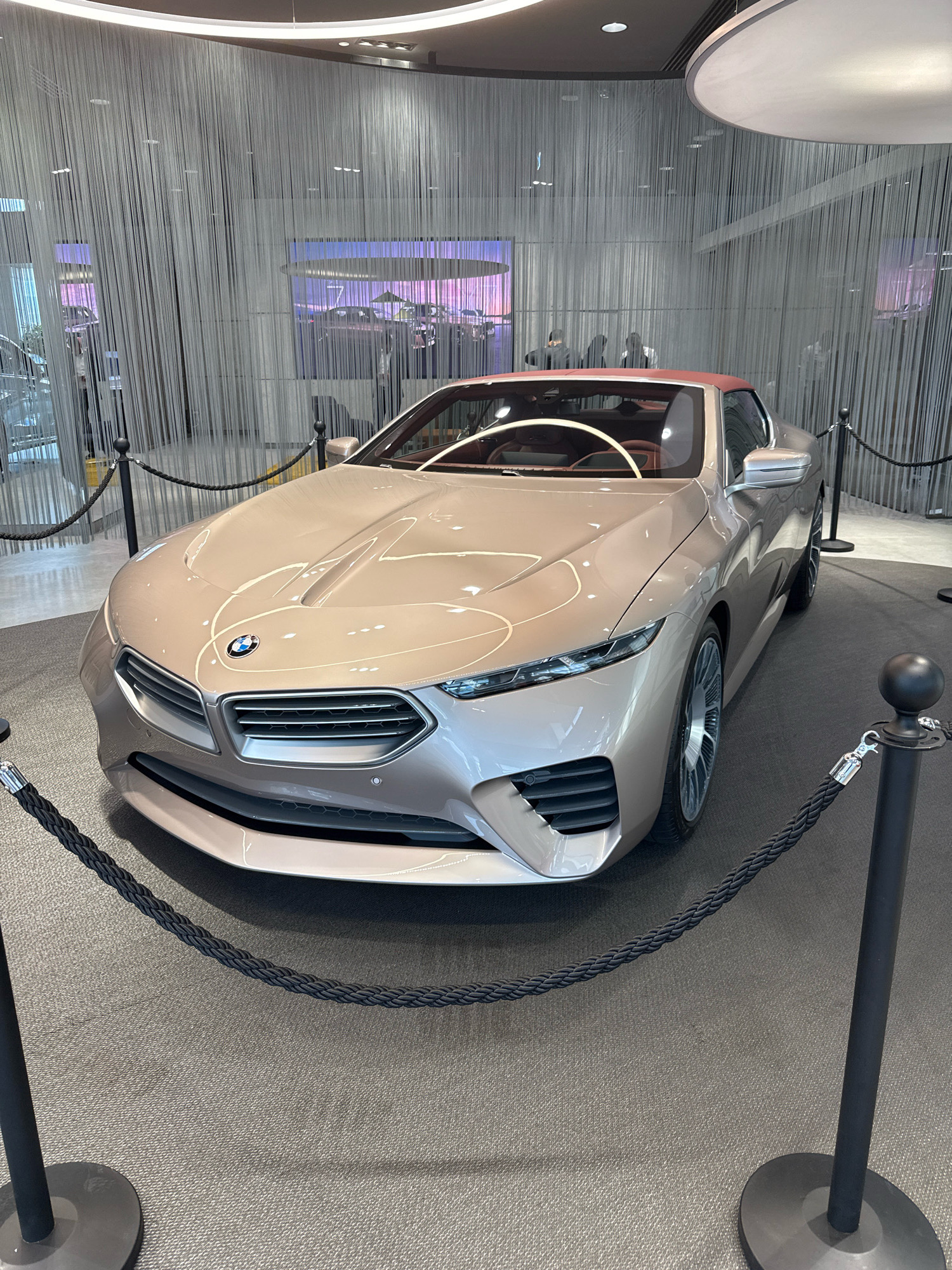 In Dubai, United Arab Emirates, a champagne-colored convertible concept car, identified as a BMW, is displayed in a showroom. The car is positioned on a grey carpeted floor, with black stanchions and ropes creating a boundary around it. The background features a striking curtain of numerous thin, vertical white strands, behind which are illuminated screens showing abstract colorful patterns and what appear to be silhouettes of people. The ceiling is visible with modern lighting fixtures. The car's interior, with its tan leather seats and white steering wheel, is partially visible. The overall environment suggests a sophisticated and modern exhibition space, likely within a high-end dealership or a dedicated automotive event venue in Dubai.