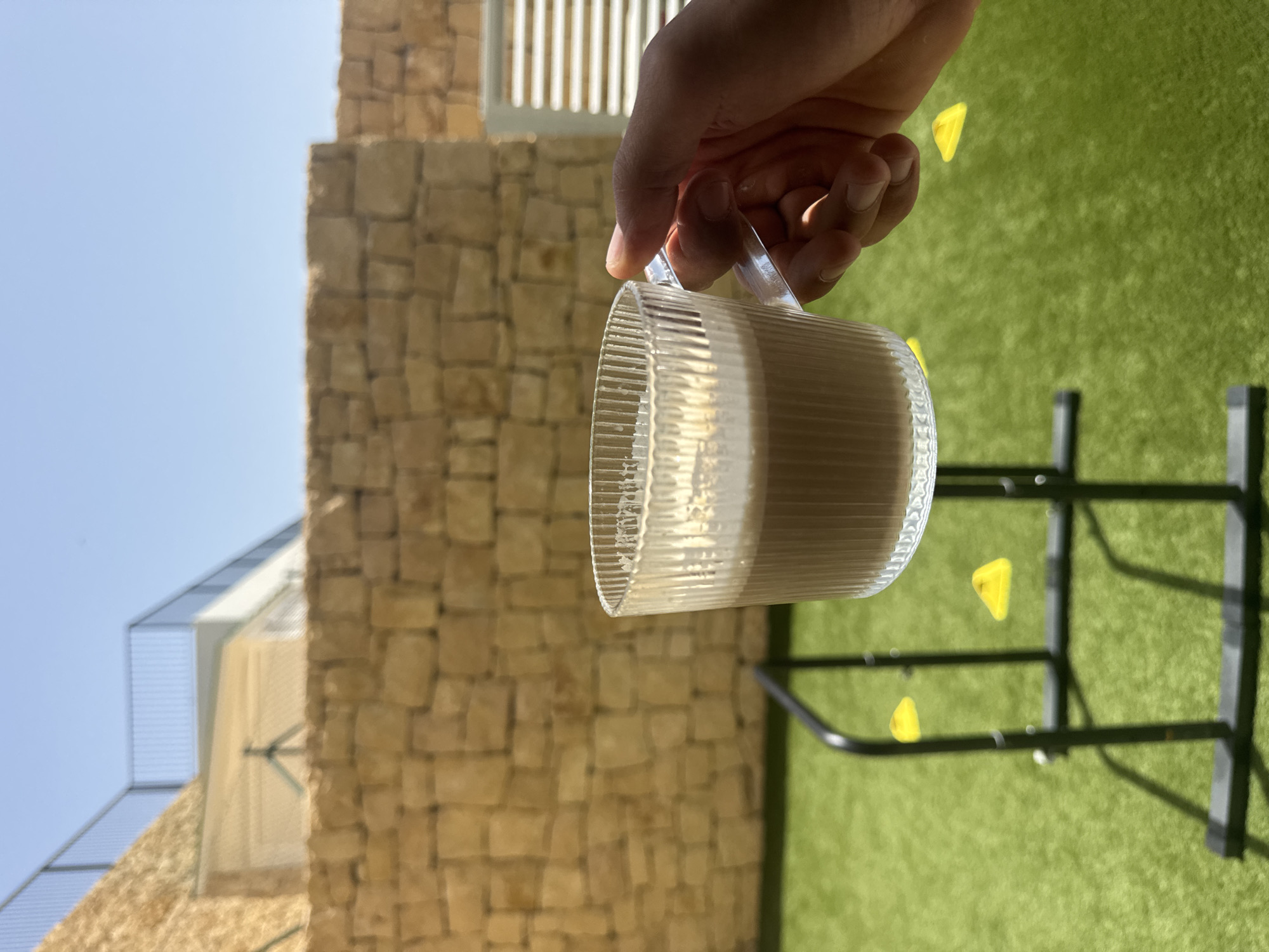 A hand holds a clear, fluted glass cup filled with a light brown beverage, likely coffee or a milkshake, with foam on top. The cup is positioned in the foreground, slightly to the right of the frame, against a blurred background. The background features a textured stone wall, a bright green artificial lawn, and a blue sky. A black metal structure, possibly a piece of outdoor gym equipment, is visible on the lawn. Yellow triangular markers are scattered on the lawn. A portion of a building with a white facade and a railing is visible above the stone wall. The scene appears to be an outdoor setting in Finestrat, Spain.