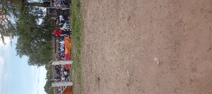 The scene shows a dirt field in Kitengela, Kenya, located at coordinates -1.47 latitude and 36.959 longitude. A group of people are sitting and standing on a grassy area, facing a speaker dressed in a red shirt. They are seated in front of an orange cloth draped over a wooden platform. The field is relatively empty and the grass is growing. Behind the people is a large tree in the middle ground, and in the background is a line of trees. The sky is cloudy.