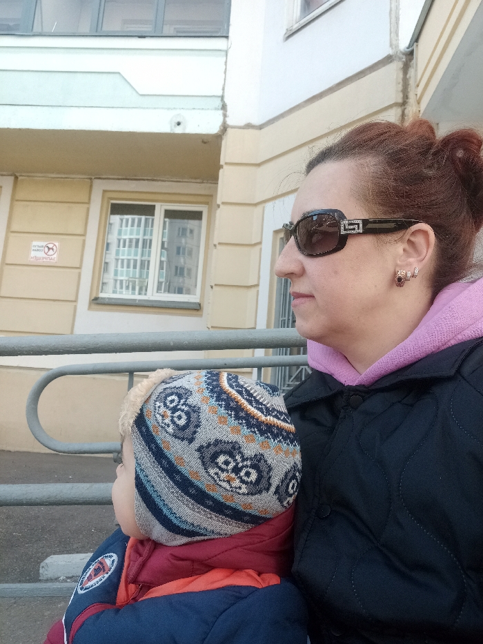 An adult female and a young child are depicted outdoors in the city of Serpukhov, Russia. The adult female, with reddish-brown hair tied back, is viewed in profile facing right. She wears dark sunglasses with prominent silver-colored, possibly rhinestone-encrusted, Greek key pattern temples, a light pink hooded sweatshirt, and a dark, possibly quilted, jacket. Small earrings with reddish stones are visible on her right ear. To her left, partially obscured, is a young child viewed in profile facing left. The child wears a multi-colored knitted winter hat with a patterned design (possibly owls) and faux fur trim, along with a red and blue winter jacket. A light-colored metal handrail extends horizontally across the middle ground. Behind the subjects, a multi-story building with light yellow walls and white trim is visible. A white, rectangular sign with a red "no dogs" symbol and Cyrillic text ("ВЫГУЛ СОБАК ЗАПРЕЩЁН" - Dog walking is prohibited) is affixed to the building's facade. A white-framed window on the building reflects a grid pattern from another structure. The scene is illuminated by natural light.