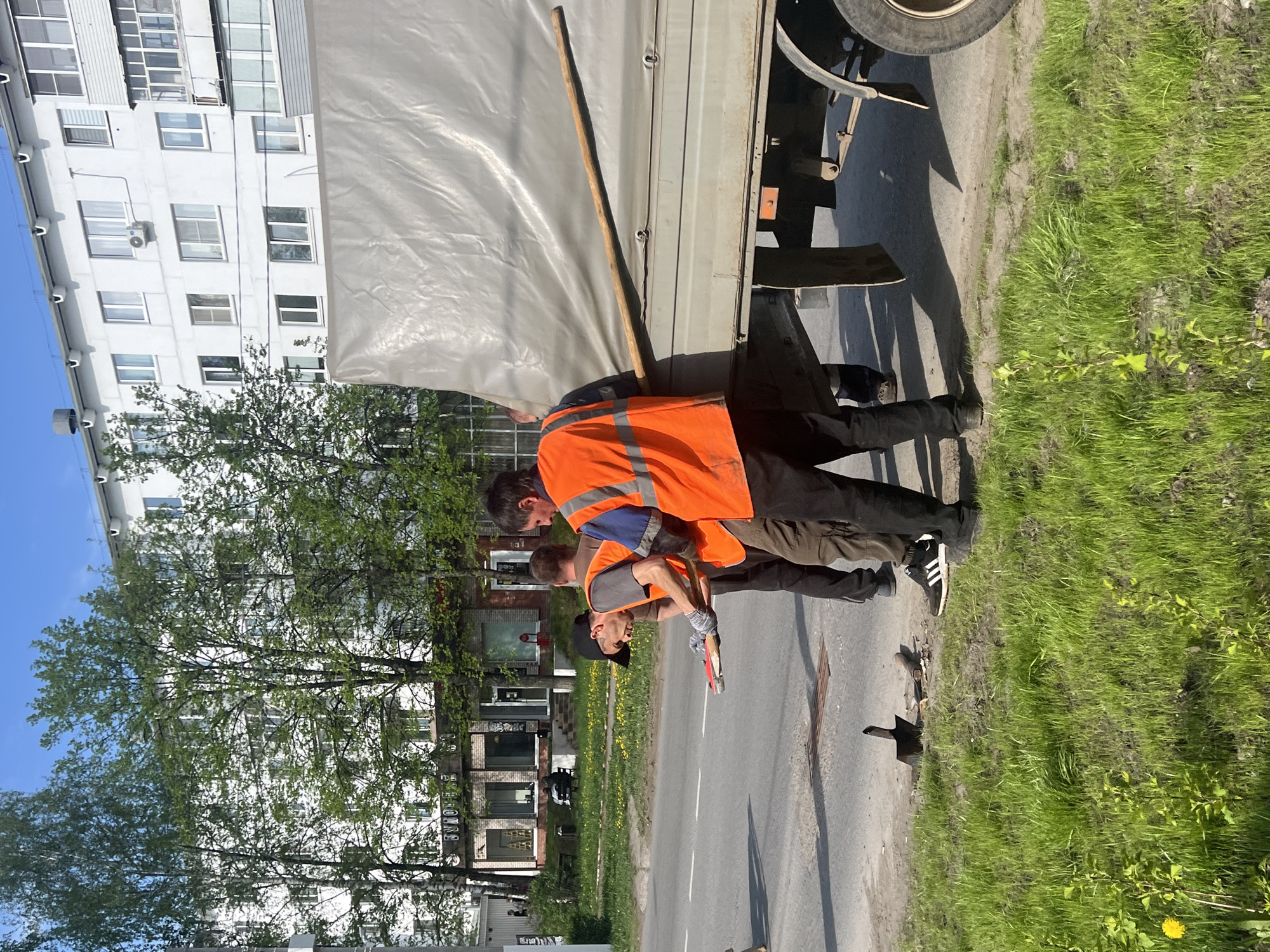 Two men stand beside a truck on a street in Chaikovsky, Russia. One man wears an orange safety vest and the other a gray shirt. The second man holds a red and silver object, possibly a tool. A building with multiple windows is visible in the background, along with trees and grass. A section of road is visible, with some discoloration and shadows.