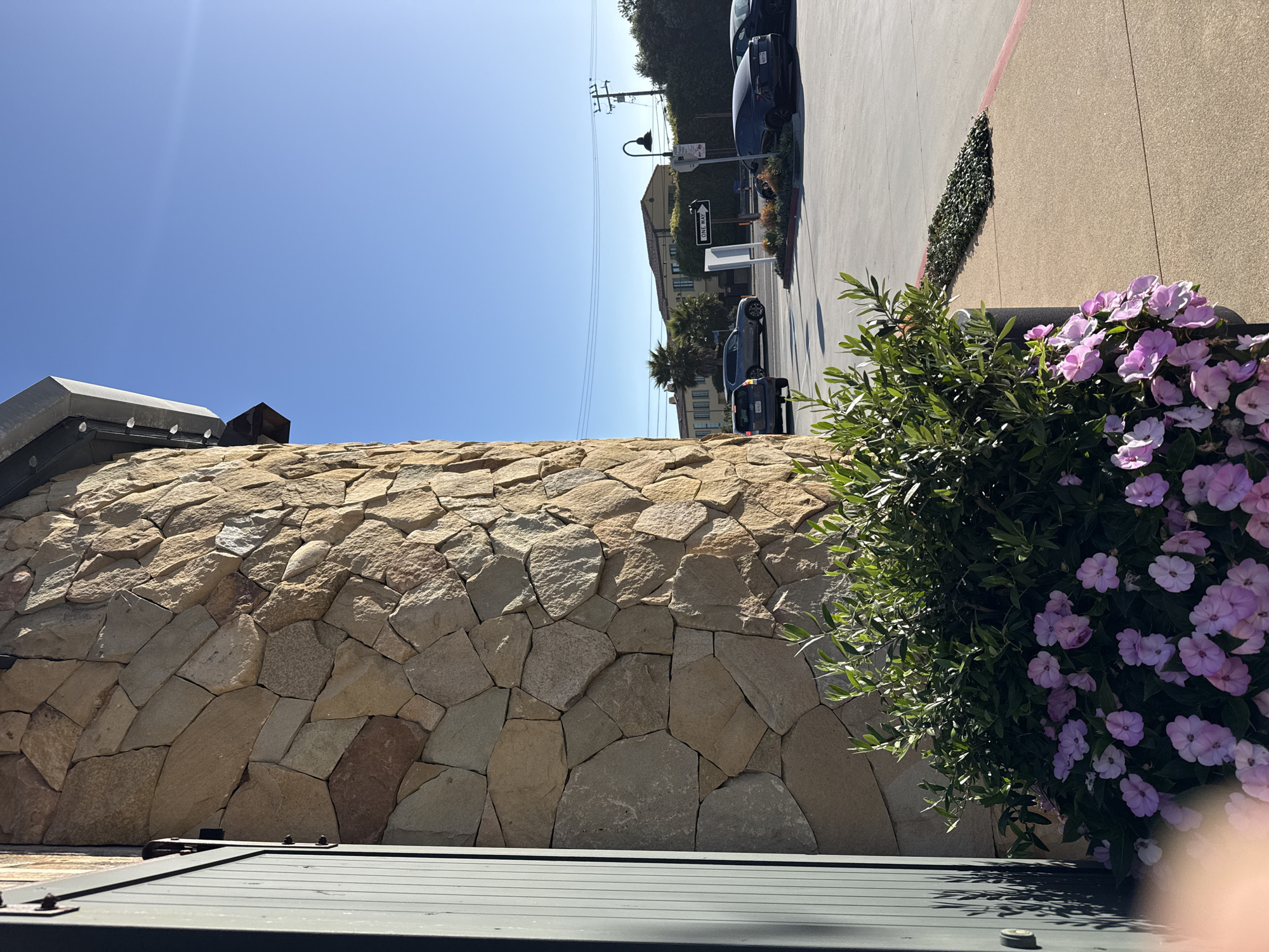 In Malibu, California, a stone-clad building stands to the left of the frame, its rough-hewn stones catching the sunlight. Adjacent to it, a planter overflows with lush green foliage and a profusion of delicate pink flowers. Beyond the building, a paved area is visible, with several cars parked. A "ONE WAY" sign points to the right, indicating traffic direction. In the background, buildings and palm trees suggest a commercial or residential area. The sky is a clear, bright blue.
