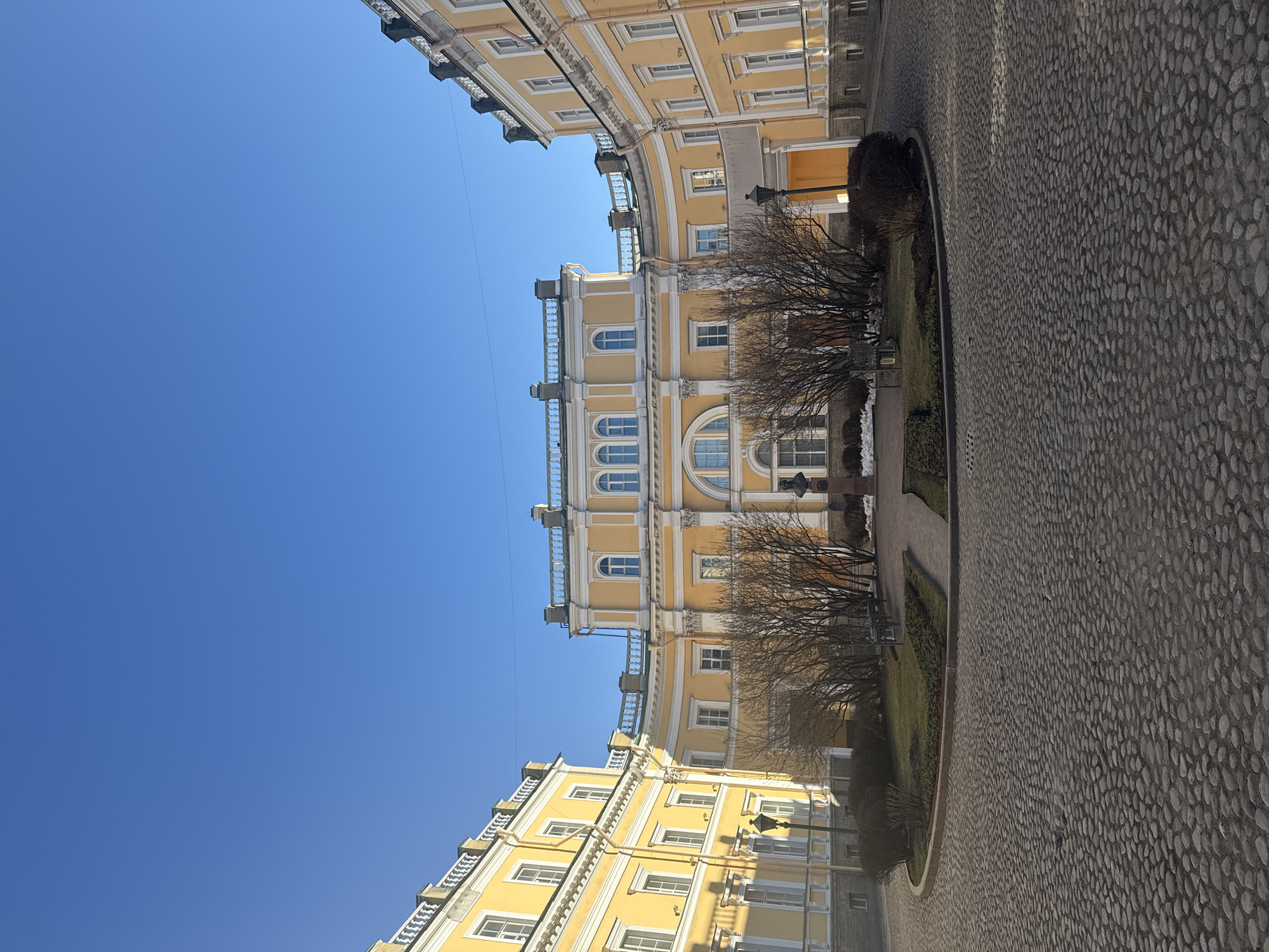 The image shows a courtyard in Saint Petersburg, Russia, framed by the yellow facades of classical buildings. The courtyard surface is paved with cobblestones, and a landscaped area with bare trees and a walkway sits in the center. The sky is a clear blue, providing a bright contrast to the buildings. There are no people visible in the scene. The architecture is characterized by neoclassical details, including white window frames, cornices, and railings. A few street lamps are visible, suggesting the area is accessible to pedestrians.