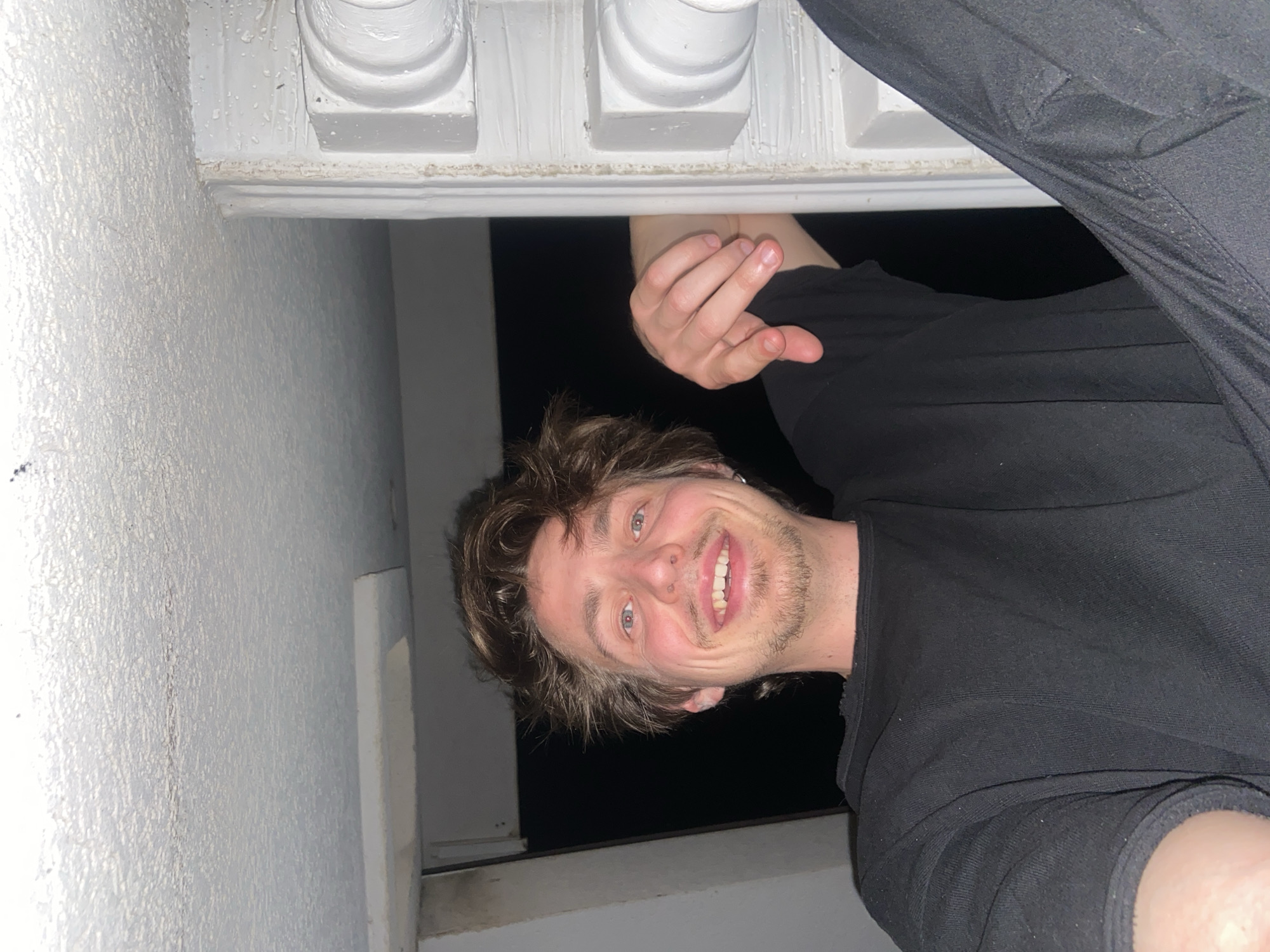 A Caucasian male is upside down and smiling at the camera. He has medium-length brown hair, blue eyes, and a light beard. He is wearing a black long-sleeved shirt and black pants. His left hand is reaching up towards a white, ornate structure above him, possibly a balcony railing. The background is a dark opening, potentially a window or doorway. To the left of the man is a textured white wall. The image appears to be taken in the dark. The location is uncertain and cannot be determined from the image without more information, so the mention of Šušanj, Montenegro is not supported by the visual evidence.