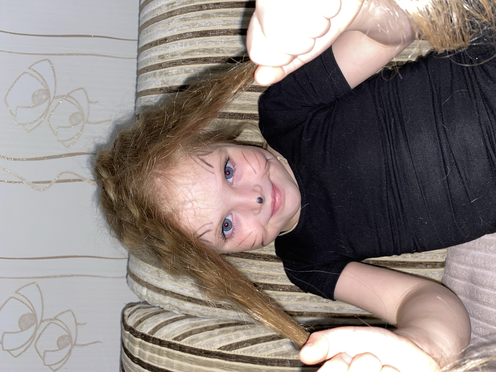 A young child with red hair and blue eyes is lying on a striped sofa, wearing a black top. The child has drawn whiskers and a nose on their face. The child is holding strands of hair in both hands. The sofa has a striped pattern. The wall behind the sofa is white with a floral pattern. The image was likely taken indoors. There is no information available in the image to suggest that the image was taken in Votkinsk, Russia.