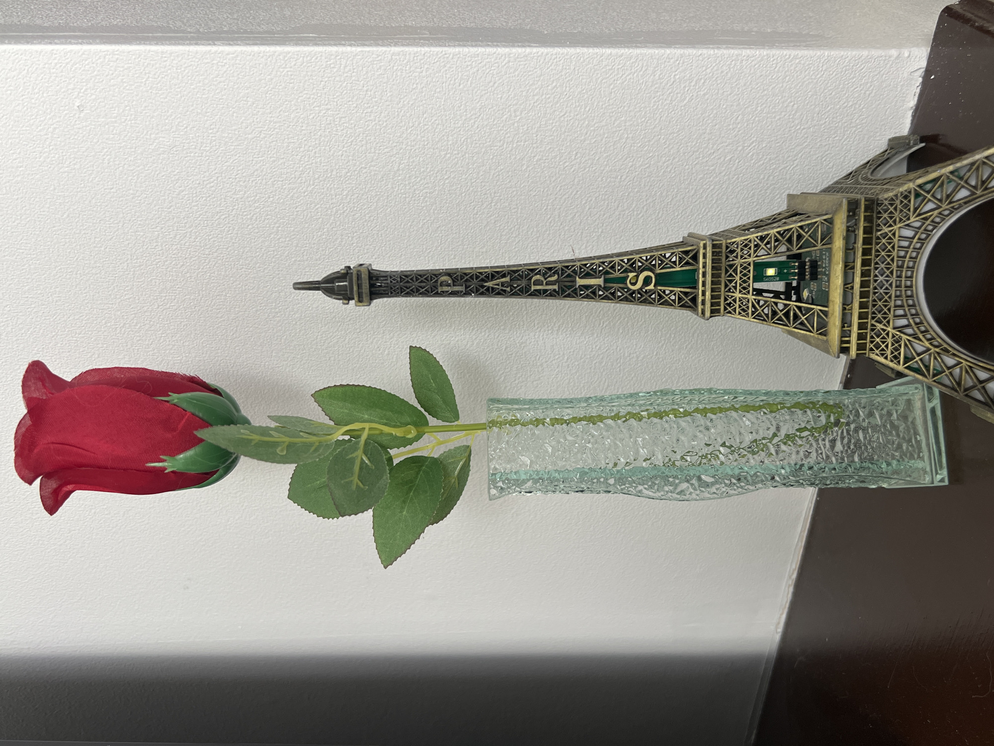 A red artificial rose in a clear, textured glass vase lies next to a miniature Eiffel Tower souvenir on a surface. The surface appears to be adjacent to a wall, which is white. The darker, possibly wooden, surface suggests the display is inside, possibly on a shelf or table. The Eiffel Tower souvenir has "PARIS" inscribed on it. The scene does not provide information about Gorleston-on-Sea, United Kingdom.