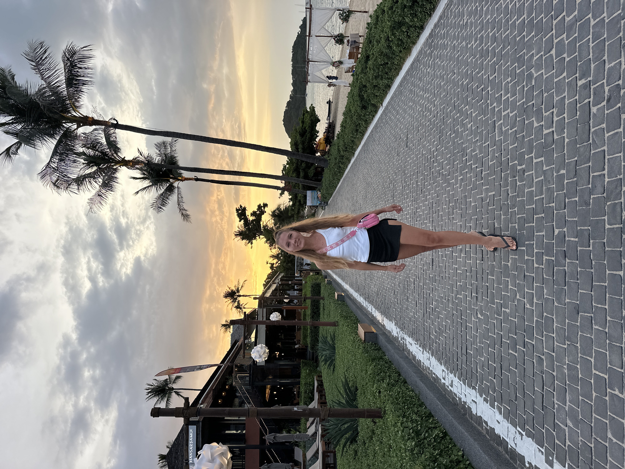 The image depicts a woman walking on a paved walkway towards the camera. She is wearing a white tank top, a black skirt, and black flip-flops, carrying a pink purse over her shoulder. The walkway is bordered by lush green bushes on the left and a sandy beach with palm trees and the ocean on the right. In the background, there are buildings, including one with the sign "HANSAR SAMUI" visible, suggesting the location is in Koh Samui, Thailand. The sky is filled with dramatic clouds, illuminated by a warm sunset glow. The scene appears to be a resort or beachfront area.