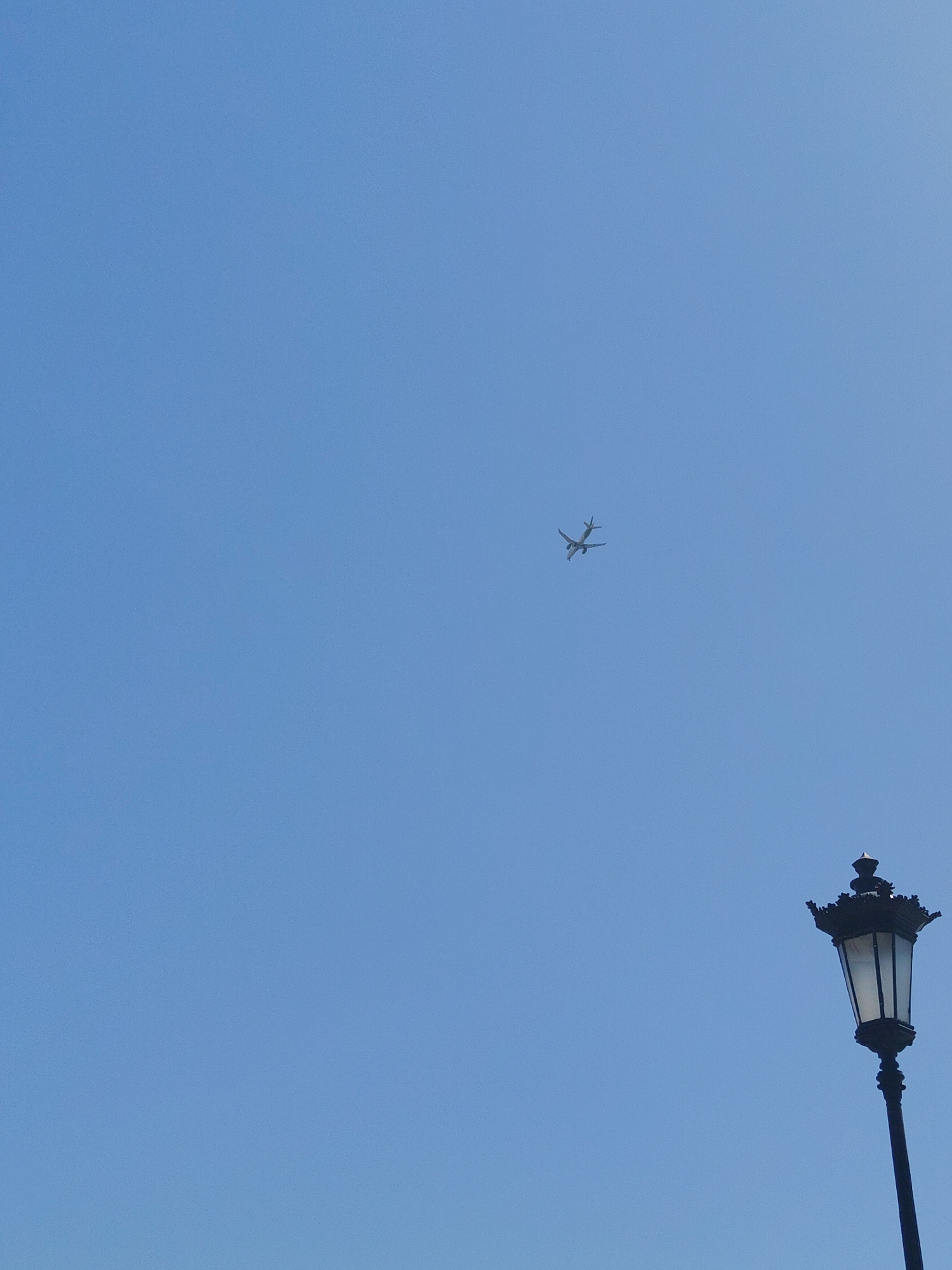 An airplane is visible in the upper right quadrant of the image, flying against a clear blue sky. In the lower right quadrant, a vintage-style lamppost with a decorative top is positioned diagonally, extending upwards. The lamppost appears to be made of black metal. The sky occupies the majority of the frame and is a uniform, cloudless blue. The scene is devoid of any people or other discernible activity. The location is the Thessaloniki Municipal Unit, Greece.