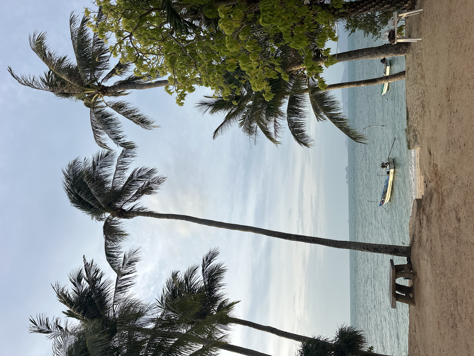 The image depicts a serene beach scene on Ko Samui District, Thailand. In the foreground, a sandy beach meets the calm turquoise ocean. A rustic wooden table and bench sit on the sand, inviting relaxation. Two small boats are anchored in the shallow water, their presence suggesting a tranquil fishing or leisure spot. Tall palm trees frame the scene, their fronds swaying gently in the breeze, adding to the tropical ambiance. The sky is overcast, with soft clouds diffusing the sunlight, creating a peaceful, subdued atmosphere. In the background, the faint outlines of islands can be seen on the horizon. The overall impression is one of a quiet, idyllic beach, perfect for unwinding.