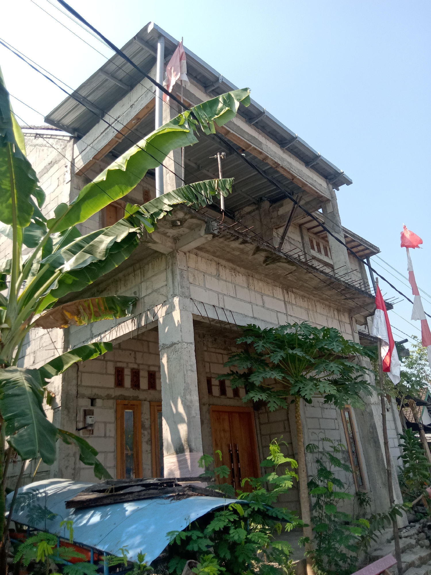 In Stiaji, Indonesia, a three-story unfinished concrete block house stands under a clear blue sky. The structure's exterior is rough, with exposed concrete and rebar visible on the upper floors. Two Indonesian flags, red and white, are displayed on poles, fluttering gently. Banana trees with large green leaves frame the left side of the house, and a smaller papaya tree grows near the entrance. The front of the house features wooden doors and windows, some of which are recessed into the concrete structure. In the foreground, a blue tarp covers an unseen object, and dense green foliage obscures the lower portion of the scene. Power lines crisscross overhead, and a meter box is mounted on the exterior wall of the house. The overall impression is of a residence under construction, with elements of tropical vegetation and national symbols.