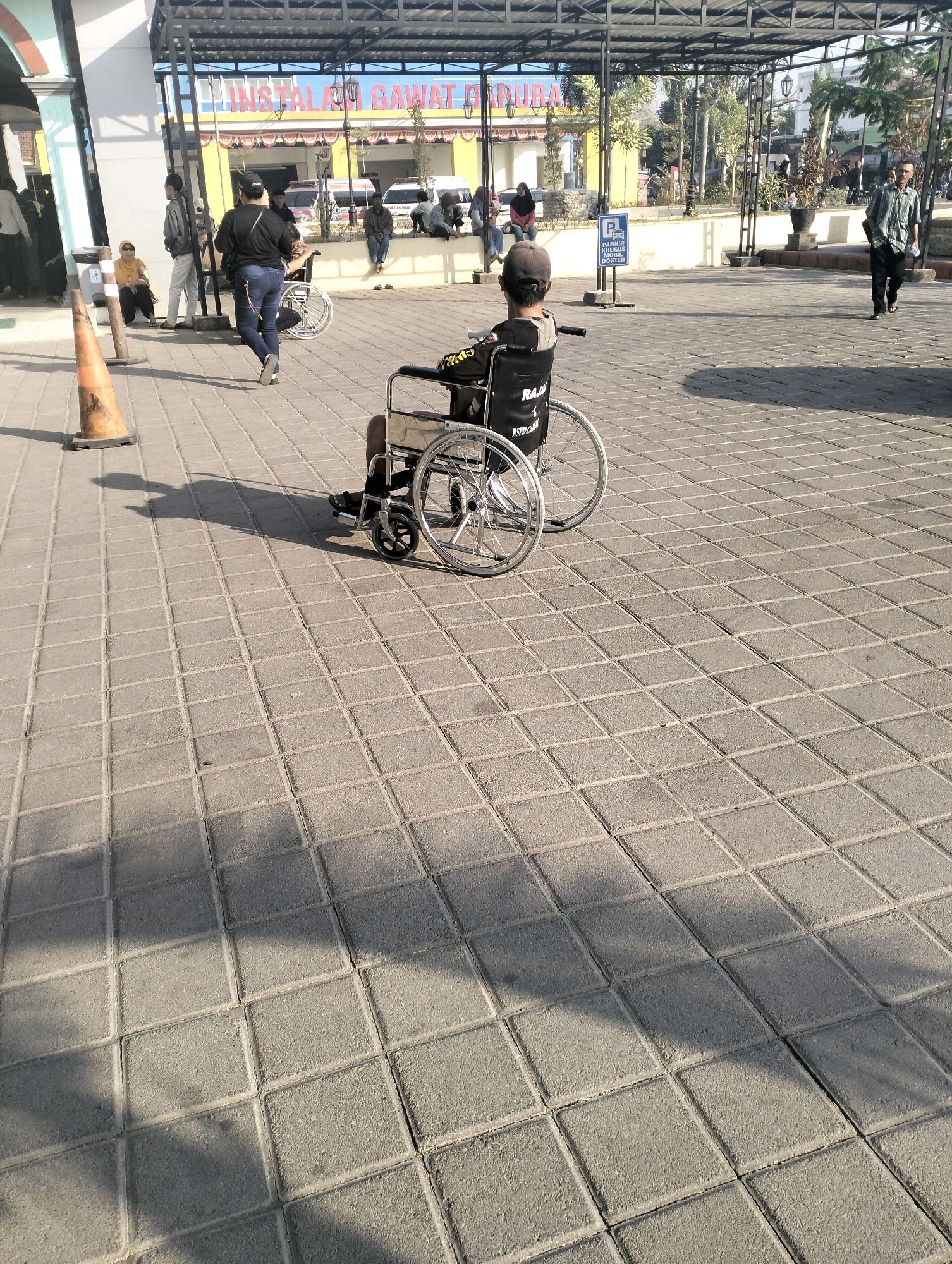 In the city of Bangunsari, Indonesia, the image captures a sunny daytime scene on a paved outdoor area. A person is seated in a wheelchair, facing away from the camera, with their back to the viewer. The wheelchair has large spoked rear wheels and smaller front wheels. To the left of the wheelchair, an orange traffic cone is visible on the paving stones. In the background, a building with a covered walkway and signage is present. Several other individuals are in the background, some walking, others sitting. The ground is a grid of square paving stones, with shadows from overhead structures and people falling across the surface. The overall impression is of a public space, possibly near a medical facility given the signage.
