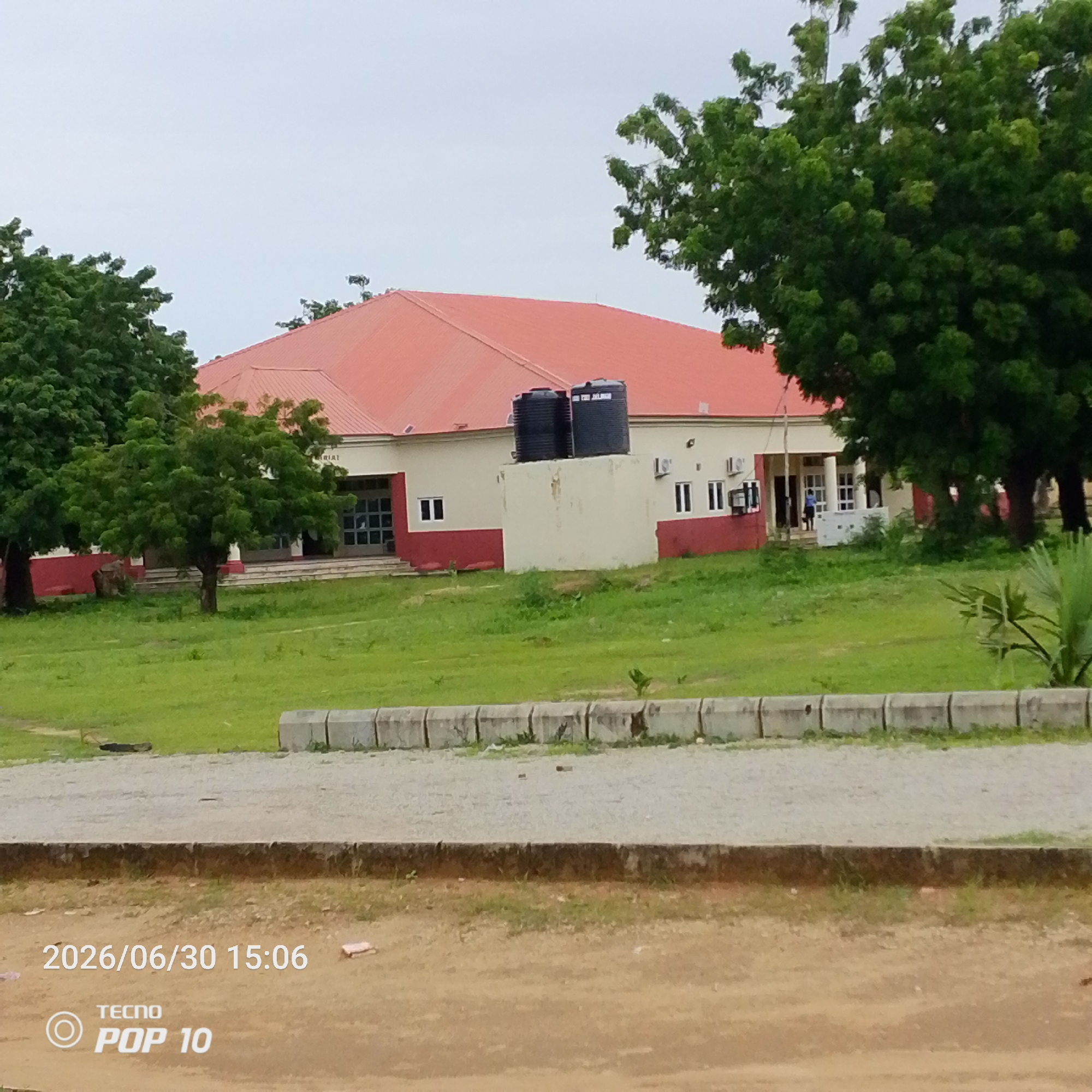 The image shows a single-story building with a prominent red roof and white walls, accented with a maroon lower section. Two large black water tanks are visible on the roof of a smaller cuboid structure attached to the main building. The building appears to be a public facility, possibly a school or community center, with several windows and entrances. A few trees, with lush green foliage, surround the building, casting shadows on the manicured lawn. In the foreground, a paved pathway separates the lawn from a dirt area where a timestamp and phone model are visible. The timestamp reads "2026/06/30 15:06", and the phone model is "TECNO POP 10". The sky is overcast, suggesting a cloudy or possibly rainy day. The setting appears to be Jalingo, Nigeria. There is no specific activity or event taking place; it is a static scene of a building and its surroundings. The overall mood is calm and ordinary.