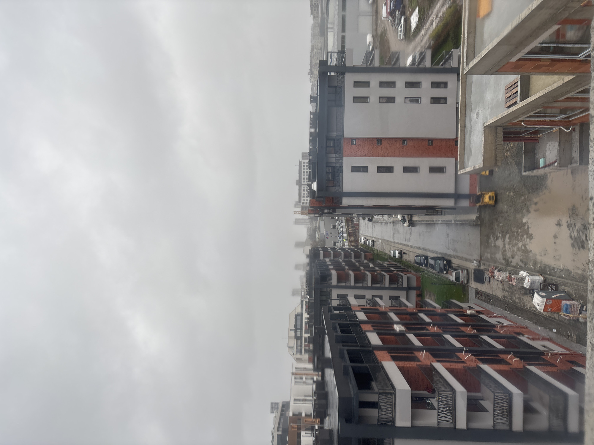 The image presents an aerial view of a cityscape, likely captured on an overcast day. The location is Burgas, Bulgaria, indicated by architectural elements and overall urban planning.
Dominating the view are several modern apartment buildings with brick and concrete facades, some still under construction. Interspersed between buildings is a street populated with parked cars. Construction equipment, including a small yellow vehicle, is visible. The sky is gray and cloudy.