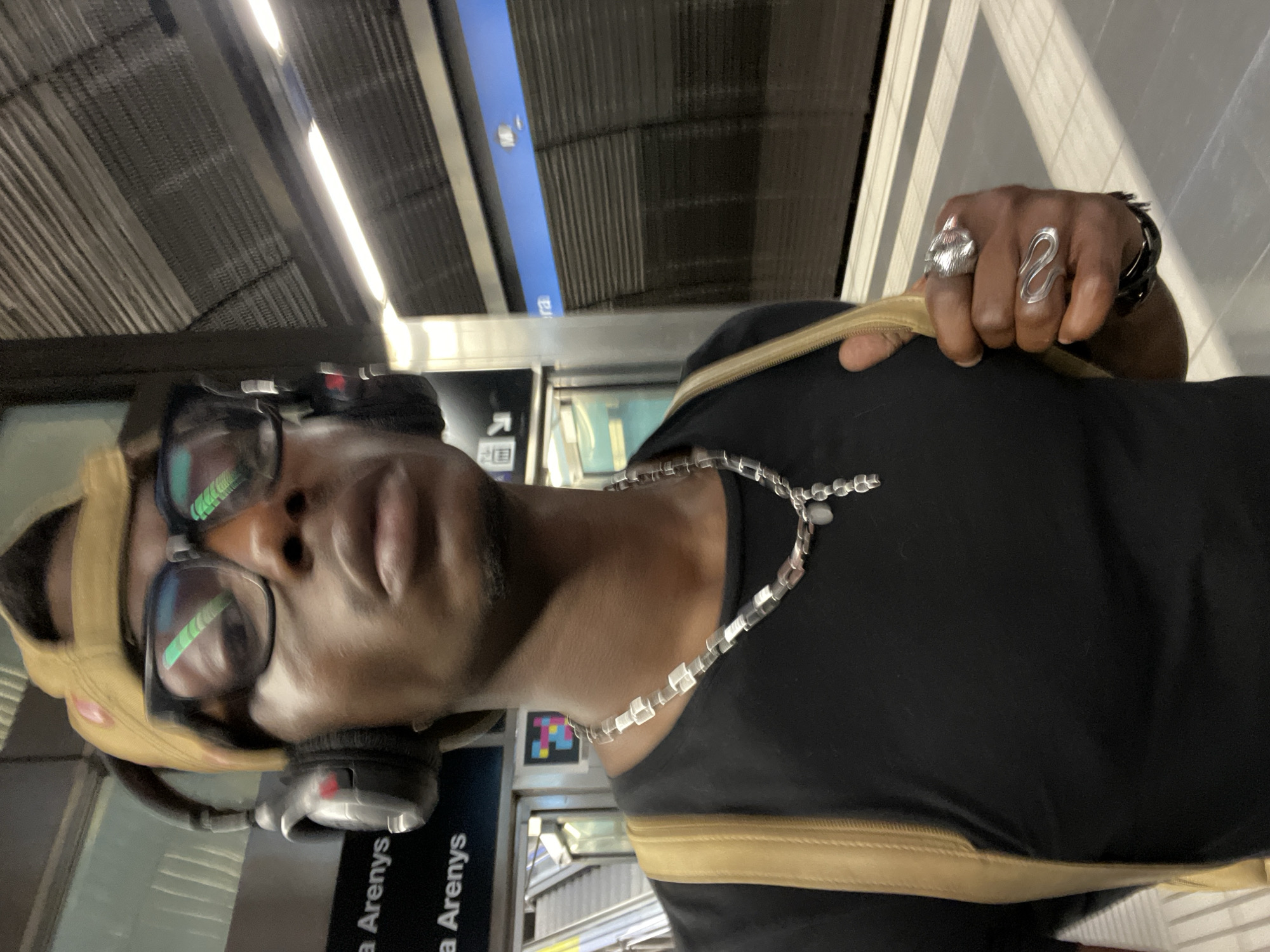 The image features a person in a public transport setting, likely a metro or train station, captured from a low angle. The subject is an individual with dark skin, wearing a black t-shirt, a tan/khaki baseball cap (worn backward), and large over-ear headphones. They wear dark-rimmed glasses that prominently reflect bright green and white linear artificial lights. A silver-colored necklace, composed of cuboid and round links, is visible around their neck. On their right hand, which holds a tan/khaki bag strap, are two distinct rings: one large and ornate, possibly depicting an animal head, and another with a sleek, wavy design. A dark wristband is also on their right wrist.
The background reveals the station's interior, characterized by metallic corrugated ceiling or wall panels and bright linear artificial lighting. Several signs are visible: a prominent black sign with white text reading "a Arenys" (repeated), and a blue sign with partially obscured text, possibly "era," and a white arrow symbol. The architectural elements and signage suggest a modern transport environment. The person appears to be posing casually, with a neutral expression, indicating they are either traveling or waiting. Given the context of Barcelona, the "Arenys" sign points to a destination within the local rail network (e.g., Rodalies de Catalunya).