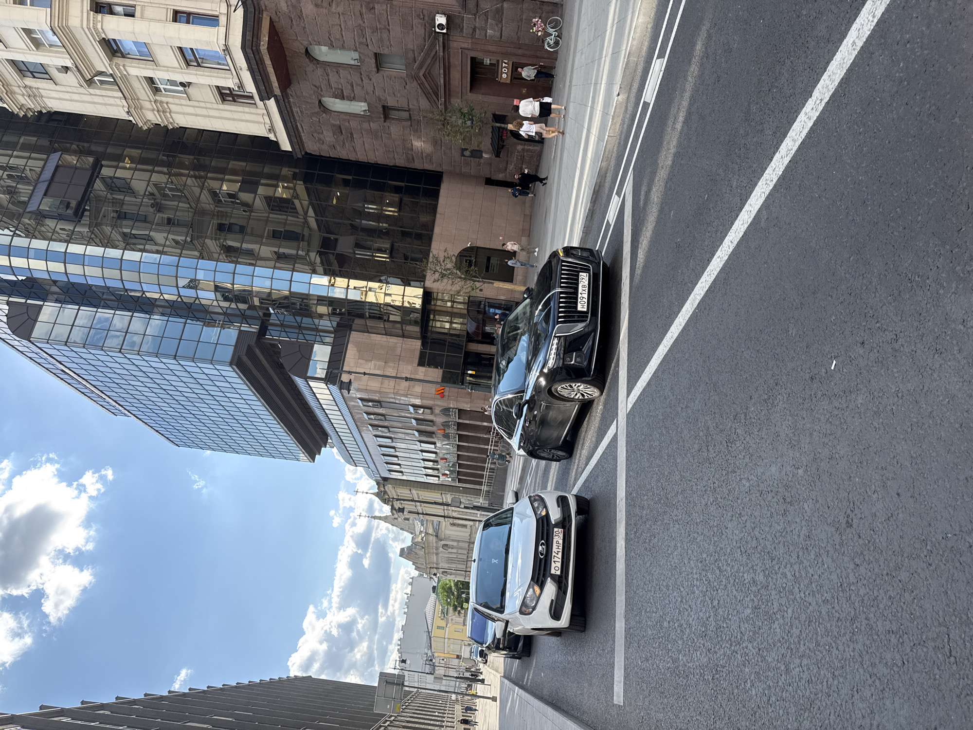 The image depicts a street scene in Moscow, Russia, on a sunny day with scattered clouds. In the foreground, a black luxury sedan is driving on an asphalt road. The car's license plate reads "H091XB 797". To the left of the sedan, a white sedan is also visible, with the license plate "O 174HP 30". Further back, a line of cars, including a black SUV and another white sedan, are stopped at an intersection.
The background features modern and historic architecture. A tall, glass-facade building dominates the right side of the frame, reflecting the sky and surrounding structures. Adjacent to it is a stone building with arched windows. Across the street, older buildings with ornate facades and church steeples are visible. Several pedestrians are walking on the sidewalk, some near shop entrances. A bicycle decorated with flowers is parked on the sidewalk near a building entrance. The overall environment suggests a busy urban setting in Moscow.