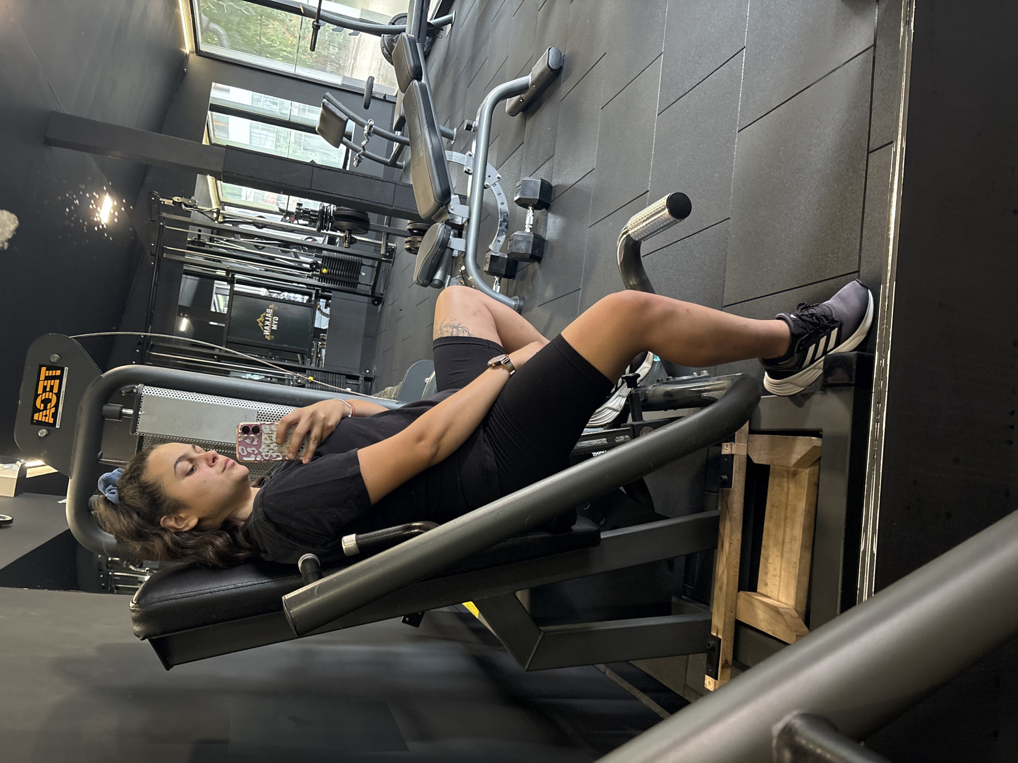 A young woman with dark hair pulled into a messy bun is sitting on a weight machine in a gym. She is wearing a black t-shirt and black shorts and has a blue scrunchie in her hair. She is holding a phone in her right hand and appears to be taking a selfie. The gym has black flooring and black walls, with various weight machines and benches visible in the background. The gym is located in Troyan, Bulgaria.
