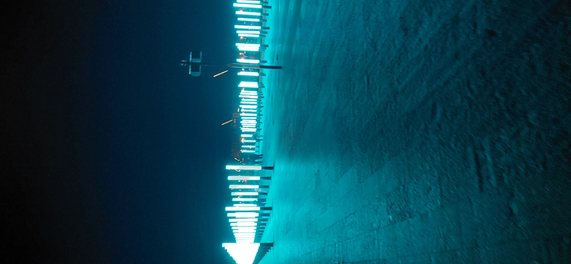 This is a nighttime photograph of an outdoor art installation featuring a long row of vertical, illuminated cylindrical lights. The lights emit a bright white light. In the background, there are several silhouetted figures of people and what appear to be animal sculptures. The ground is covered in a light dusting of snow or frost, and the overall lighting is dominated by the artificial light of the installation, casting a cool, bluish-green glow. The scene suggests a public space, possibly a park or pedestrian walkway, in Magnitogorsk, Russia, decorated for an evening event or exhibit.