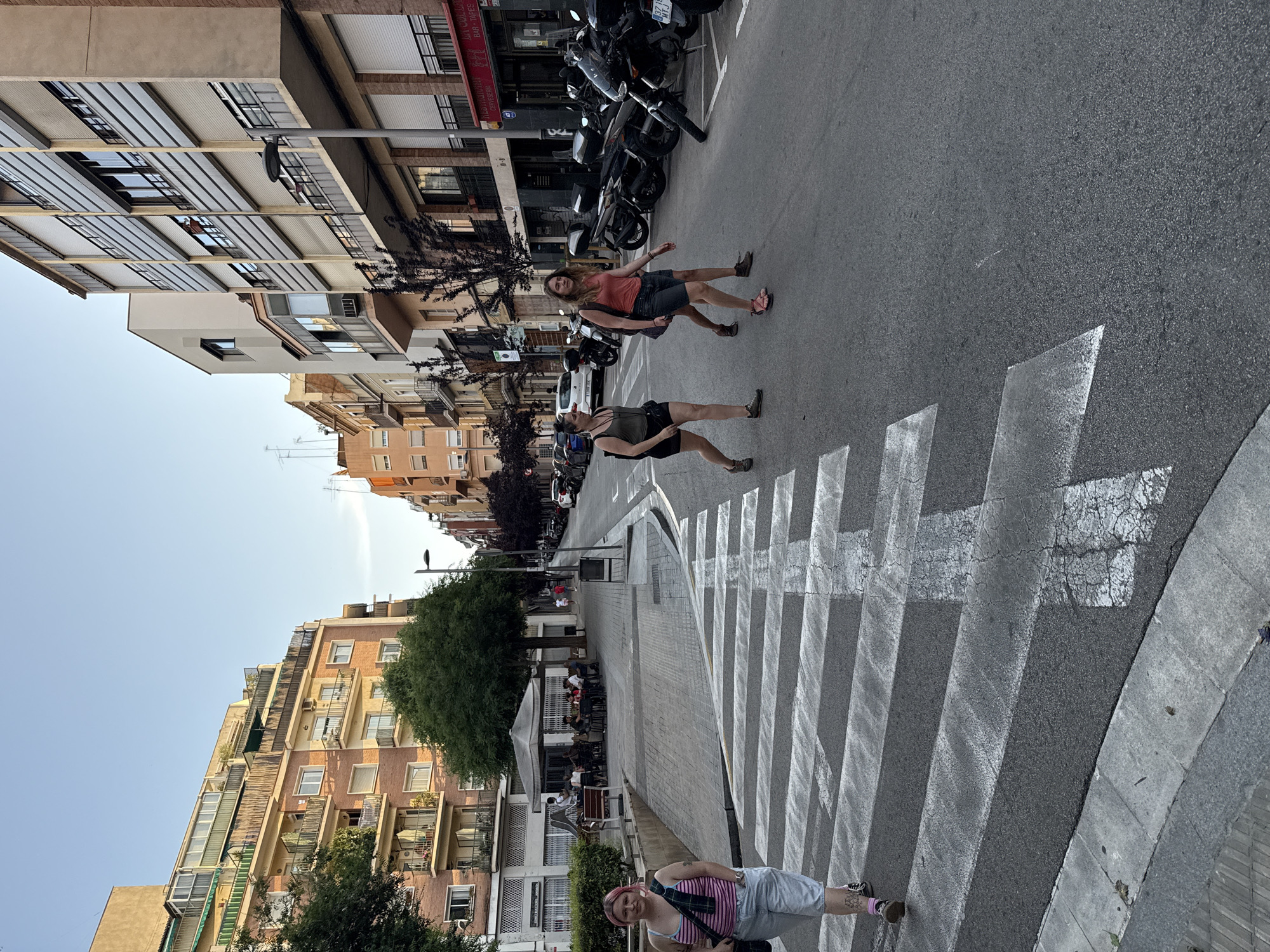 In the city of Barcelona, Spain, a pedestrian crossing is prominently featured in the foreground of a street scene. The crossing is marked with wide white stripes on asphalt. A young woman with pink hair, wearing a pink and white striped tank top, denim shorts, and black shoes, walks across the street on the left side of the frame, facing the camera. Behind her, two other women are crossing the street from right to left. The woman on the left is wearing a dark tank top and black shorts, while the woman on the right is wearing a coral-colored tank top and dark shorts. They are walking together and appear to be interacting.
The background shows a street lined with multi-story buildings, typical of urban Barcelona. Some buildings have balconies and awnings. Trees are planted along the sidewalk, and outdoor seating areas with umbrellas are visible in front of a cafe or restaurant. Several motorcycles are parked on the right side of the street. The sky is clear and blue, indicating daylight. The overall scene depicts a casual street life in a European city.