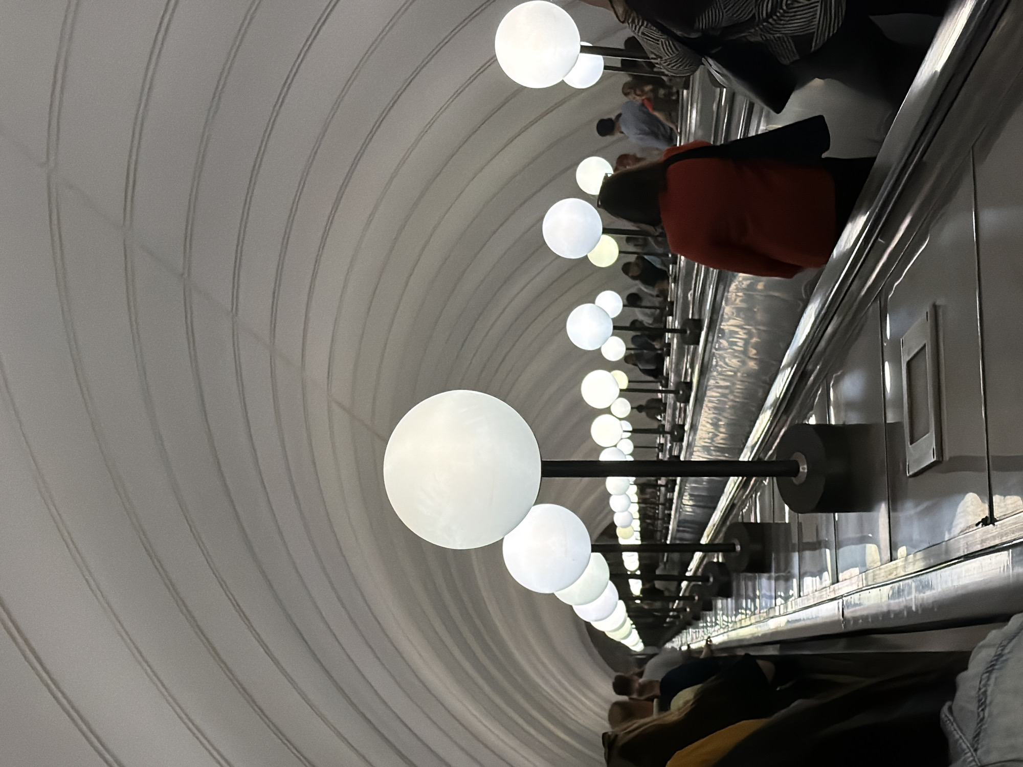 The image depicts a long, curving escalator tunnel, likely in a subway station, characterized by its arched, white ceiling with parallel grooved accents. Along the right side of the escalator, a series of large, spherical, white light fixtures on dark poles illuminate the scene. Several people are visible, either standing on or riding the escalator. The perspective is from the perspective of someone on the escalator, looking forward and slightly to the left. The escalator treads are metallic and reflective. The overall atmosphere is subdued and functional, with the prominent lighting creating a sense of depth and perspective within the tunnel.