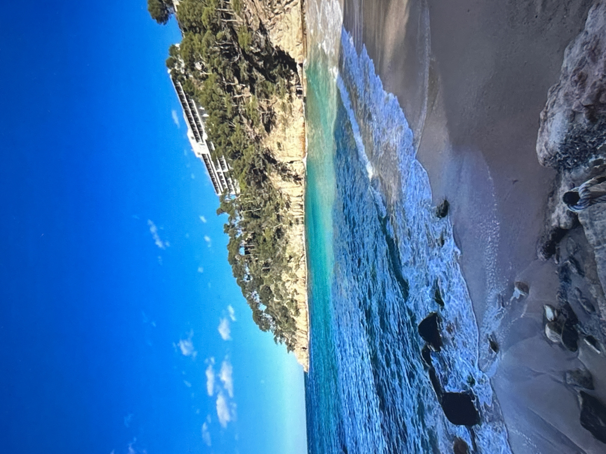 The image displays a vibrant coastal landscape featuring a beach, the sea, a cliff, and a large building under a bright blue sky.
The scene is dominated by the calm, clear blue-green waters of the sea, which meet a sandy beach along the right side of the frame. Gentle waves with white foamy crests lap at the shore. The beach itself is a mix of light-colored sand and scattered dark rocks, some of which are partially submerged in the shallow water.
Rising steeply from the back of the beach is a rugged cliff, densely covered with green trees and Mediterranean-style vegetation. Nestled into this verdant cliffside, a prominent white, multi-story building is visible, likely a hotel or resort, with numerous windows and balconies facing the sea.
Above the land and sea, the sky is a brilliant, clear blue with a few scattered, wispy white clouds. The bright illumination suggests it is daytime, likely mid-morning or afternoon, with sunny weather. There are no individuals or specific activities visible, creating a serene and undisturbed atmosphere. No text is discernible in the image.