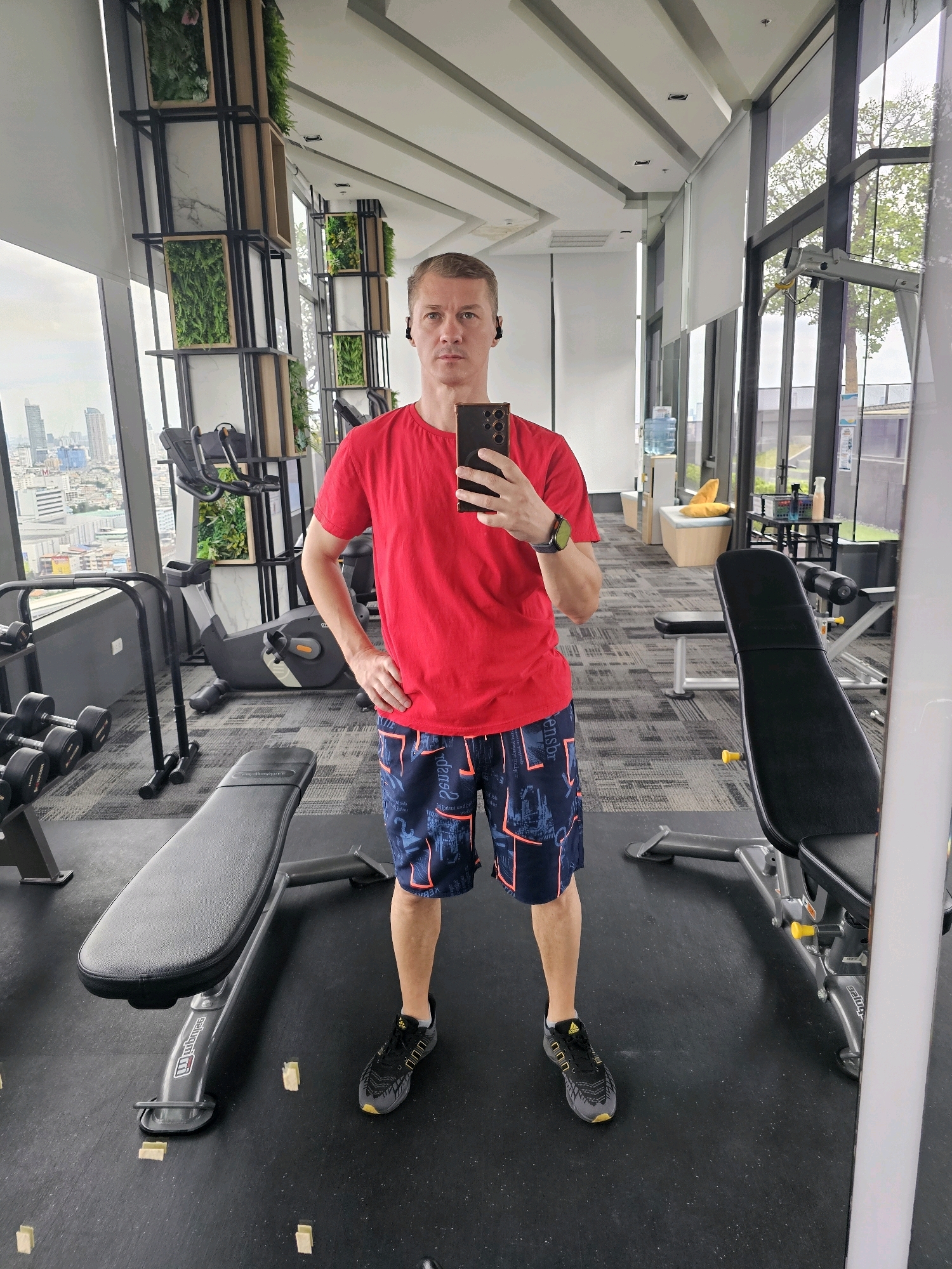 A man is taking a mirror selfie in a gym located in Bangkok, Thailand. The man is wearing a red t-shirt, dark blue shorts with an orange pattern, black and yellow athletic shoes, and black wireless earbuds. He is holding a black smartphone in his right hand and has his left hand on his hip. The gym is well-lit and features large windows that offer a view of the city. Equipment such as exercise bikes, dumbbells, and weight benches are visible in the background. Decorative vertical plant installations are mounted on pillars. The flooring is dark grey with a textured pattern.