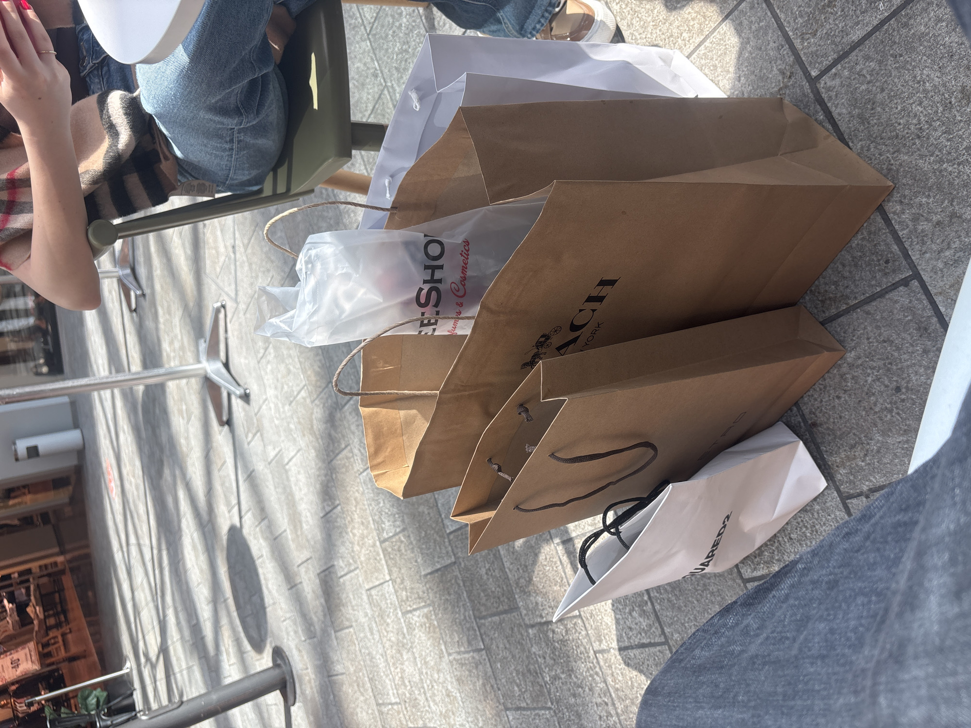The image captures an outdoor scene in Mendrisio, Switzerland, showing a collection of shopping bags placed on a light grey paved ground. A person is partially visible on the left, seated at a table, wearing blue jeans and a brown and beige plaid or striped scarf/shawl. Their hands, with red nail polish on the fingernails, are visible resting near a white tabletop. Another person's leg, clad in dark jeans, is partially visible on the far right.
The shopping bags include one large white paper bag with white handles, three brown paper bags with string handles, and a smaller white paper bag with black handles at the bottom right. A clear plastic bag is visible inside one of the brown bags, bearing the text "FREE-SHOP" and "Perfumes & Cosmetics". Another prominent brown bag features the "COACH" logo, including its carriage icon, and the words "NEW YORK". The text "SQUARED2" is partially visible on the white bag at the bottom right.
The ground is composed of light grey rectangular pavers, and strong, distinct shadows suggest bright sunlight, indicating it is likely midday or early afternoon. Metal supports for outdoor furniture are also visible on the ground, creating a setting consistent with an outdoor café or seating area where people might take a break during a shopping excursion.