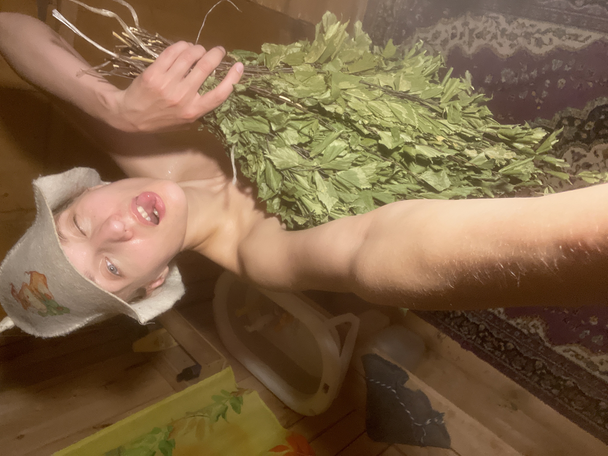 A person is lying down in a wooden sauna, wearing a felt sauna hat and holding a bundle of leafy branches. The person's mouth is open, and their tongue is sticking out. The sauna is dimly lit, with steam visible in the air. There are wooden benches and a small bucket on the floor. A patterned rug is visible in the background. The image appears to be a selfie, taken from a low angle. The lighting and steam suggest a warm and humid environment.