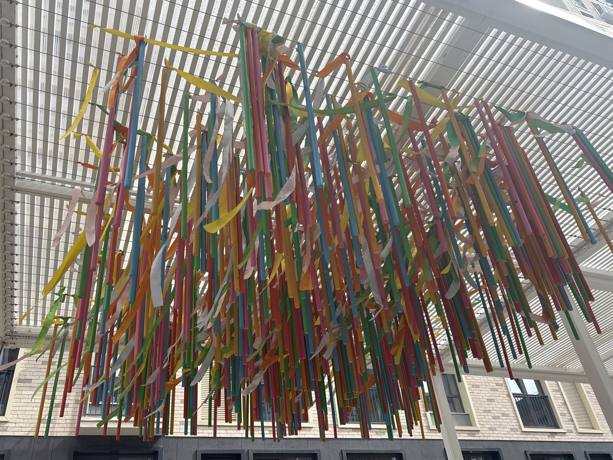 A low-angle, wide shot captures a decorative installation under a white pergola-like structure. The installation consists of numerous brightly colored wooden rods of varying lengths suspended from the pergola. Ribbons in corresponding colors are tied to the tops of the rods.
The colors of the rods and ribbons include yellow, orange, pink, red, blue, and green. The rods are densely packed, creating a visually striking effect.
In the background, several stories of a building can be seen, with brick and dark gray facade elements. The building has black-framed windows. The pergola's structure consists of parallel white beams.
The photo was taken in Izhevsk, Russia.