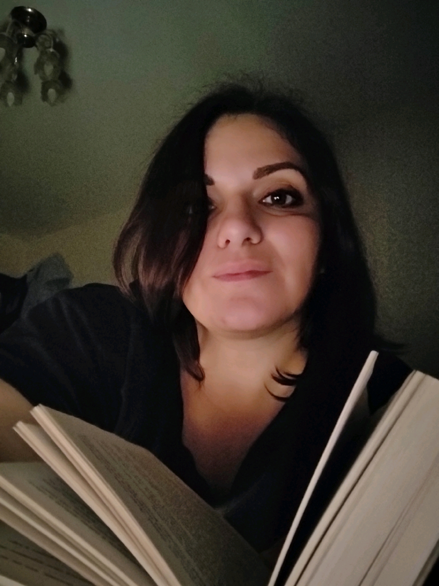 A medium shot captures a woman from the chest up, holding an open book. She has dark, shoulder-length hair and is wearing a dark-colored shirt. The lighting is dim, casting shadows and creating a subdued atmosphere. The background is a plain, textured wall in a muted green tone. The woman's face is angled towards the camera, and she appears to be looking at the book or the viewer. The scene is likely indoors, given the artificial lighting and indoor setting suggested by the wall texture. No specific location within Марковский, Russia, can be determined from the image.