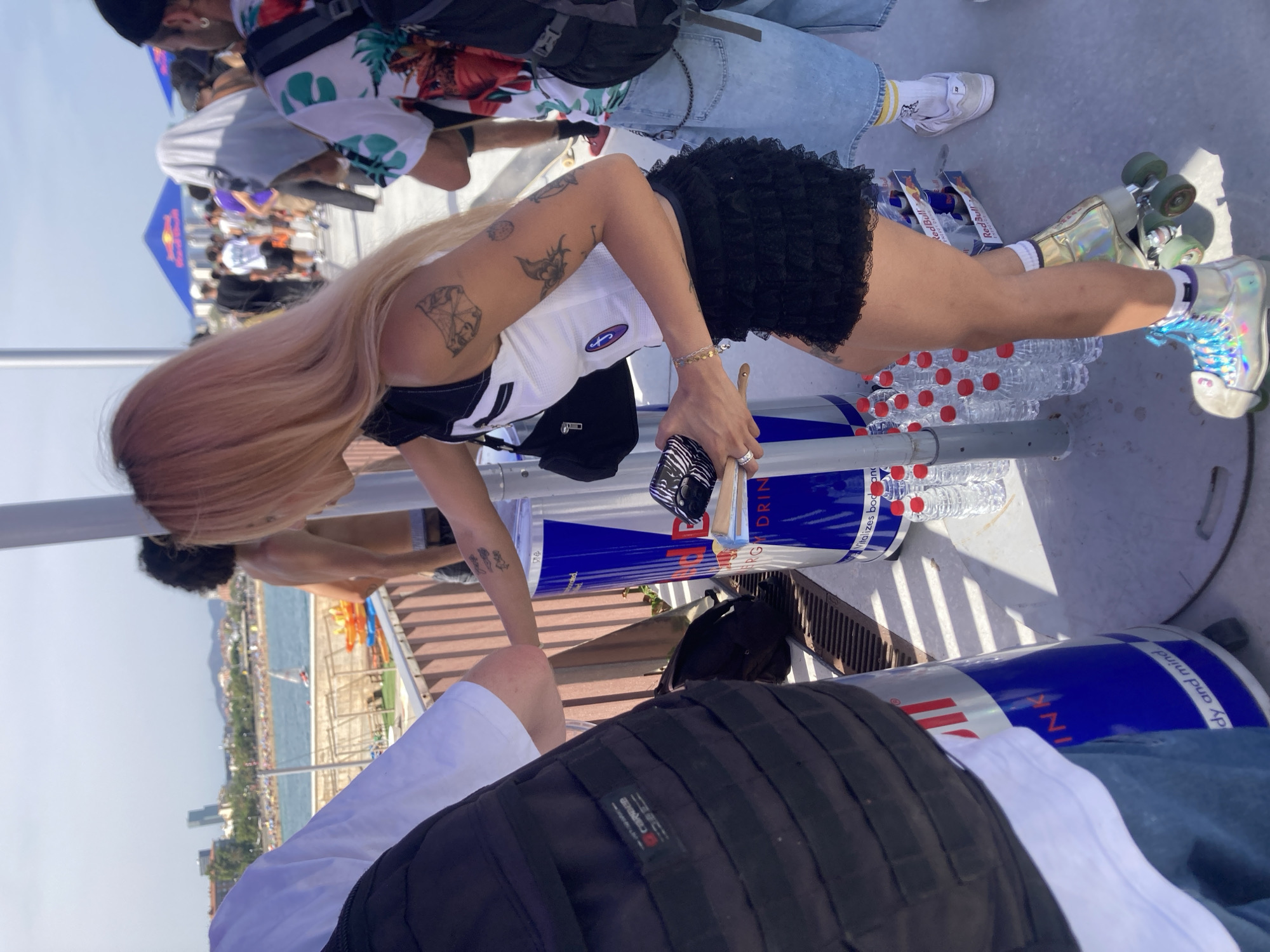 The image depicts an outdoor event near the Barceloneta beach in Barcelona, Spain. In the foreground, a young woman with blonde-pink hair, wearing a black and white cropped top and a black ruffled skirt, is standing on roller skates. She is interacting with a large Red Bull branded bin. She is holding a brush and some papers. Beside her, to the left, a person wearing a white t-shirt and a black backpack is partially visible. To the right of the woman, another person is standing, wearing a colorful patterned shirt and denim shorts. In the background, the Mediterranean Sea is visible, with several boats on the water and the cityscape of Barcelona lining the coast. People are milling about, suggesting a public gathering or event. The environment is sunny, indicated by the bright lighting and shadows.