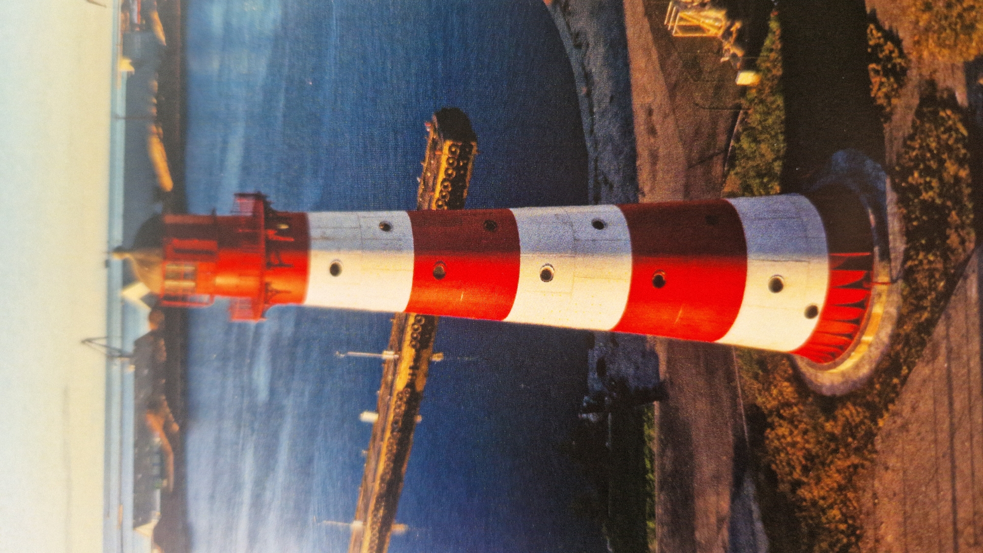 An aerial, eye-level view captures a red and white striped lighthouse standing tall in the foreground, its red lantern room capped with a dark roof. The lighthouse is positioned on a slightly elevated, grassy area with some surrounding vegetation. In the mid-ground, a calm body of water, likely the Baltic Sea, extends towards a long pier or jetty. Beyond the water, industrial structures, including cranes and buildings, are visible on the distant shoreline, suggesting a port or harbor area. The sky above is a pale, hazy blue with hints of sunlight. This scene is characteristic of the Liepaja Port in Liepaja, Latvia, a common location for such maritime infrastructure.