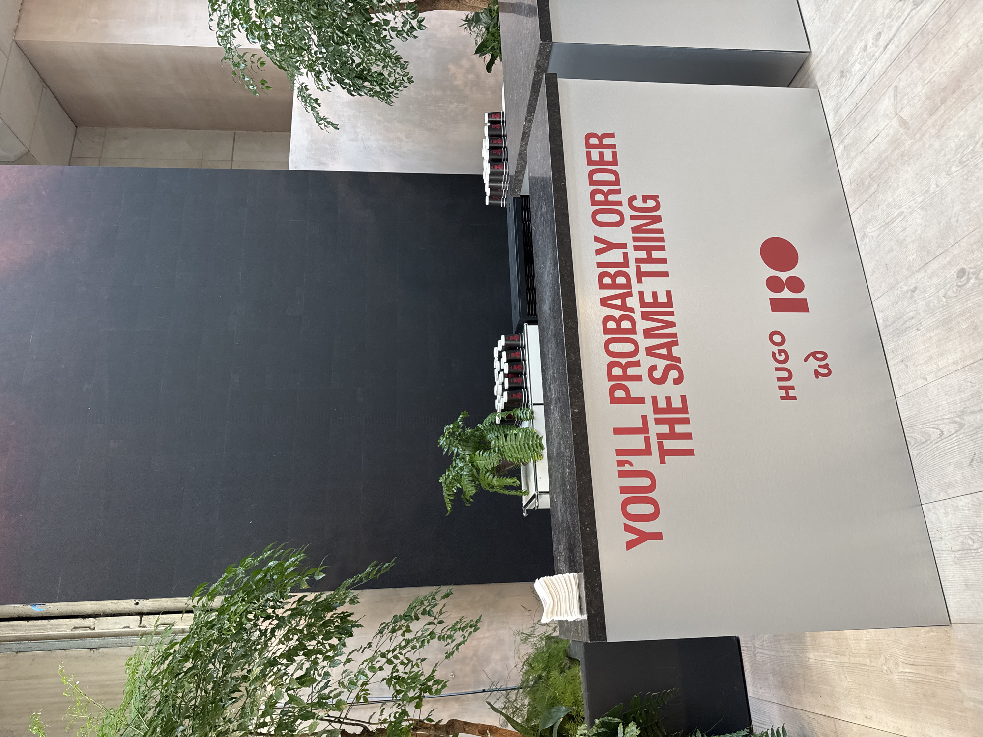 The image captures a section of a modern interior space, likely a retail or hospitality venue, in London, United Kingdom. The dominant feature is a display unit with the text "YOU'LL PROBABLY ORDER THE SAME THING" printed in red, alongside the words "HUGO" and "UD 180" also in red. The display unit is positioned on a floor with a light wood-grain pattern. Behind the display, a dark-colored wall provides a contrasting backdrop. Potted plants, including trees and ferns, are placed around the display and in the surrounding area, adding a natural element to the modern design. Several stacks of items, possibly disposable cups, are organized on the top surface of the display. A glimpse of the building's architecture reveals a concrete or plaster wall in the upper part of the frame. The scene appears to be clean and well-maintained, suggesting a contemporary and deliberate design aesthetic.
