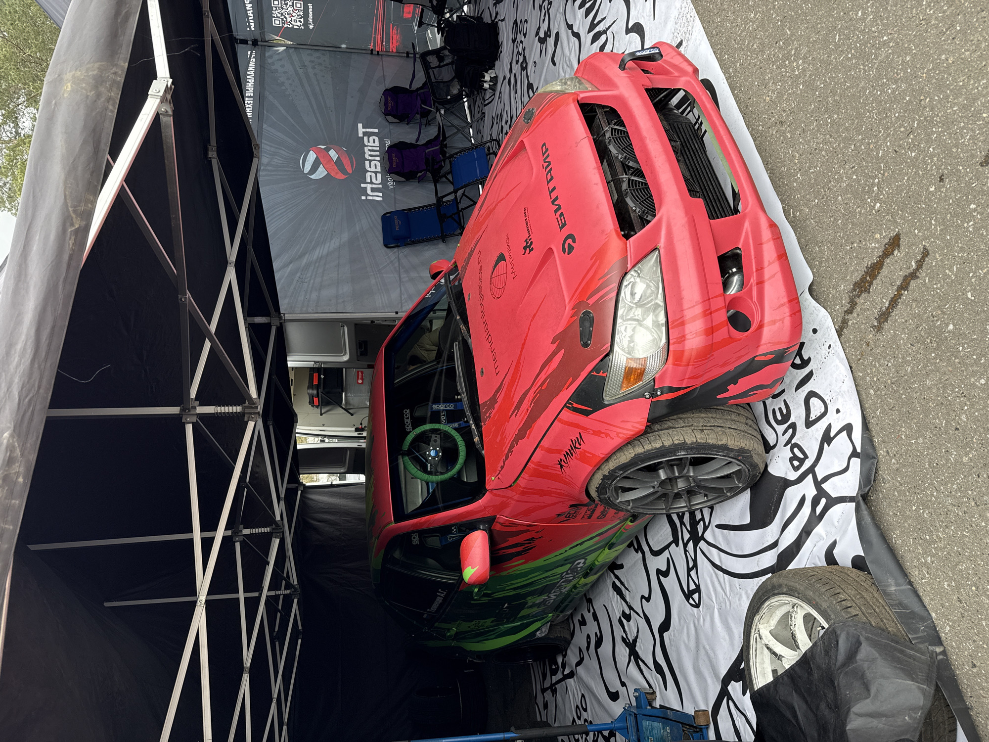 A red and green modified race car, likely a drift car, is parked under a black canopy tent in what appears to be a pit area. The car has a distinctive custom livery, red on the upper half transitioning to green on the lower half, with black accents. Its front bumper is removed, revealing the radiator and cooling fans, suggesting it is either undergoing maintenance or is prepared for enhanced cooling during a performance event. Inside the car, a bright green Sparco steering wheel is visible.
The scene depicts an outdoor motorsport event during the daytime, under soft, possibly overcast, lighting. The ground under the tent is covered with a black and white patterned mat, while outside the tent, it is asphalt. Several folding chairs and spare wheels/tires are also present within the tent setup. No people are visible.
Visible text includes:
- On the car: "БИТАЙР" (BITAIR) on the hood, "meridianlogistics.ru" accompanied by a globe logo, "Кулики" (Kuliki) near the front wheel, "Тюменцев А.Г." (Tyumentsev A.G.) on the side window, and multiple "sparco" logos (including on the steering wheel and fender).
- On a backdrop banner: A logo with intertwined 'S' shapes, and "Tamsai" (mirrored as "irL6msT" in OCR) with a QR code and mirrored text "qi.irlzomot" (likely a website when unmirrored).
- On folding chairs: "PRESENT" and "ΠΛΑΝΟ" (PLANO).
- On the ground mat: Partially visible text "BUE DIA".
The setting is in Ostrovtsy, Russia.